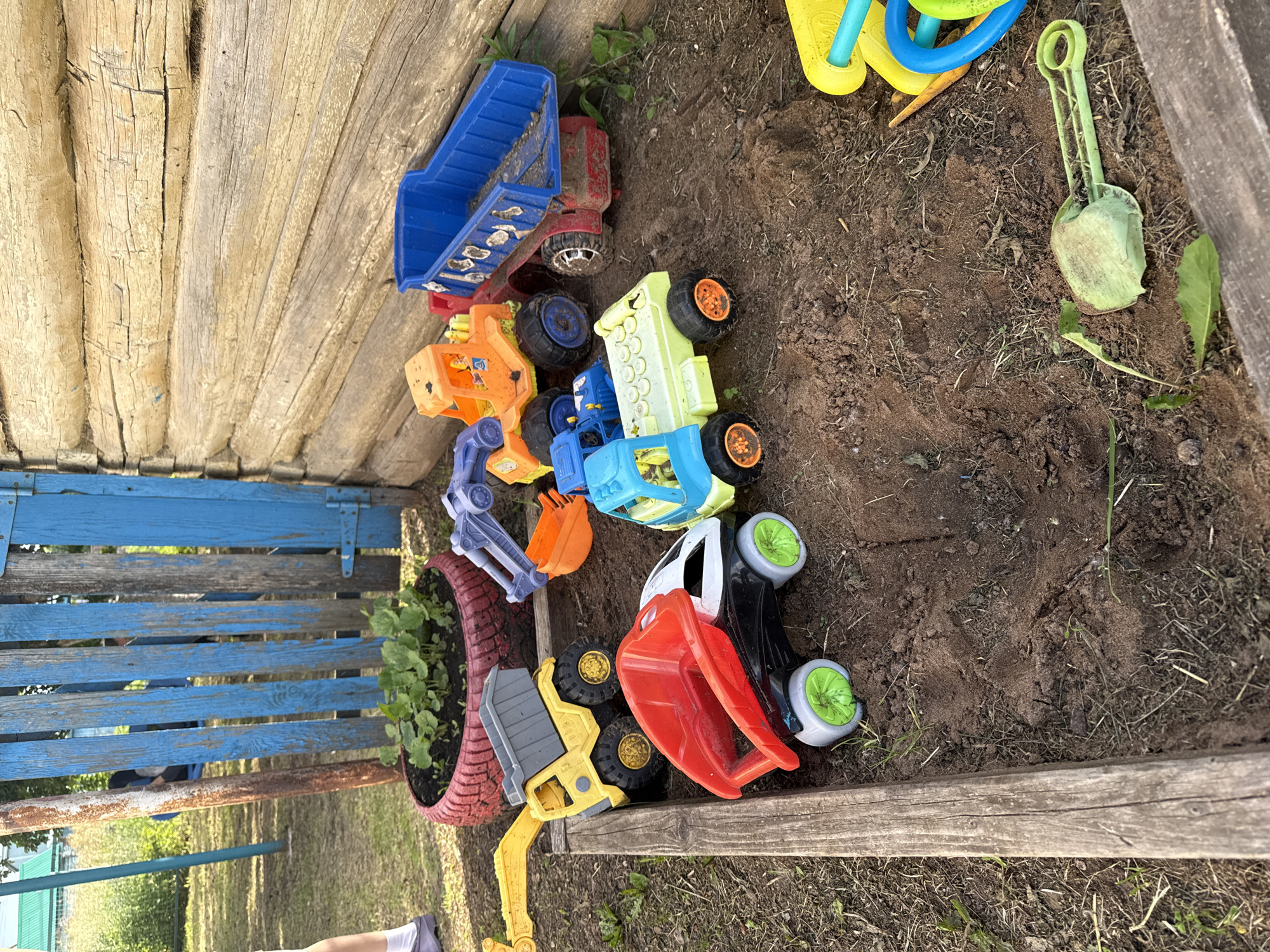 This is a medium-shot, eye-level, outdoor photograph taken in a sandbox at a property in Chuy-Atasevo, Russia. The sandbox is bordered by wooden planks and contains several toy construction vehicles, including a yellow and grey dump truck with an excavator arm, a red and black dump truck, a light green and blue dump truck, an orange and blue digger, and a blue dump truck. Scattered around the sandbox are a green plastic shovel and a yellow and blue watering can. In the background, there is a wooden fence and a small garden bed planted with seedlings in a red tire. The ground is covered in dirt and dry grass.