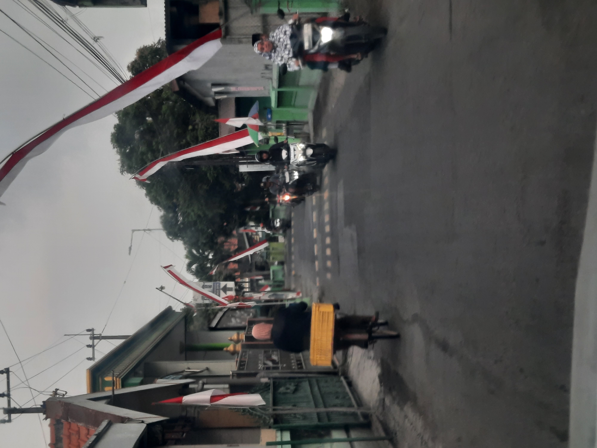 The image depicts a street scene in Banjarkemantren, Indonesia, under an overcast sky. Indonesian flags, red and white, are prominently displayed, draped along the street and affixed to buildings. Several people are visible, mostly on motorcycles, with one person riding a bicycle in the foreground. The person on the bicycle is carrying a yellow crate. The buildings on either side of the street appear to be houses or small businesses. The road is paved, and there are utility poles with wires overhead. The overall impression is of a community preparing for or celebrating an event, possibly a national holiday, given the prevalence of the flags.