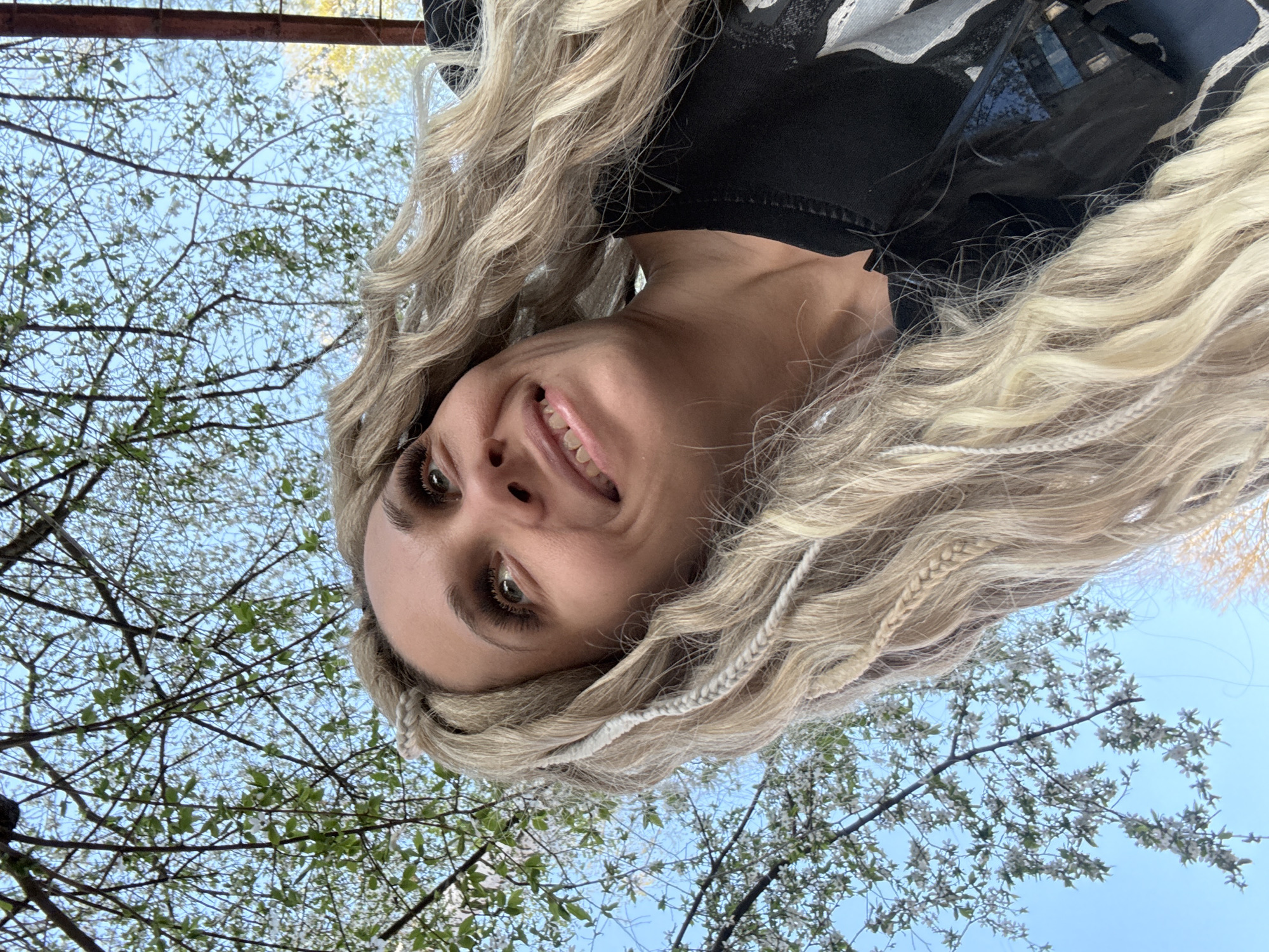 A woman with long, blonde hair smiles at the camera. She is wearing a black top and is positioned with her head tilted to the side. Behind her, tree branches with green leaves and white blossoms extend across a clear blue sky. The photograph was taken outdoors, presumably in a park-like setting, but there are no discernible landmarks to confirm the specific location within Rusanovo, Russia.
