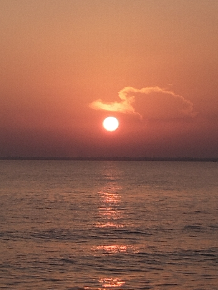 The image captures a serene sunset over the ocean. The sky is a warm blend of orange and red hues, with the sun appearing as a bright, round orb partially obscured by a cloud. The ocean reflects the sunlight, creating a shimmering path towards the viewer. The water's surface is slightly rippled, adding texture to the scene. A thin strip of land is faintly visible on the horizon.
