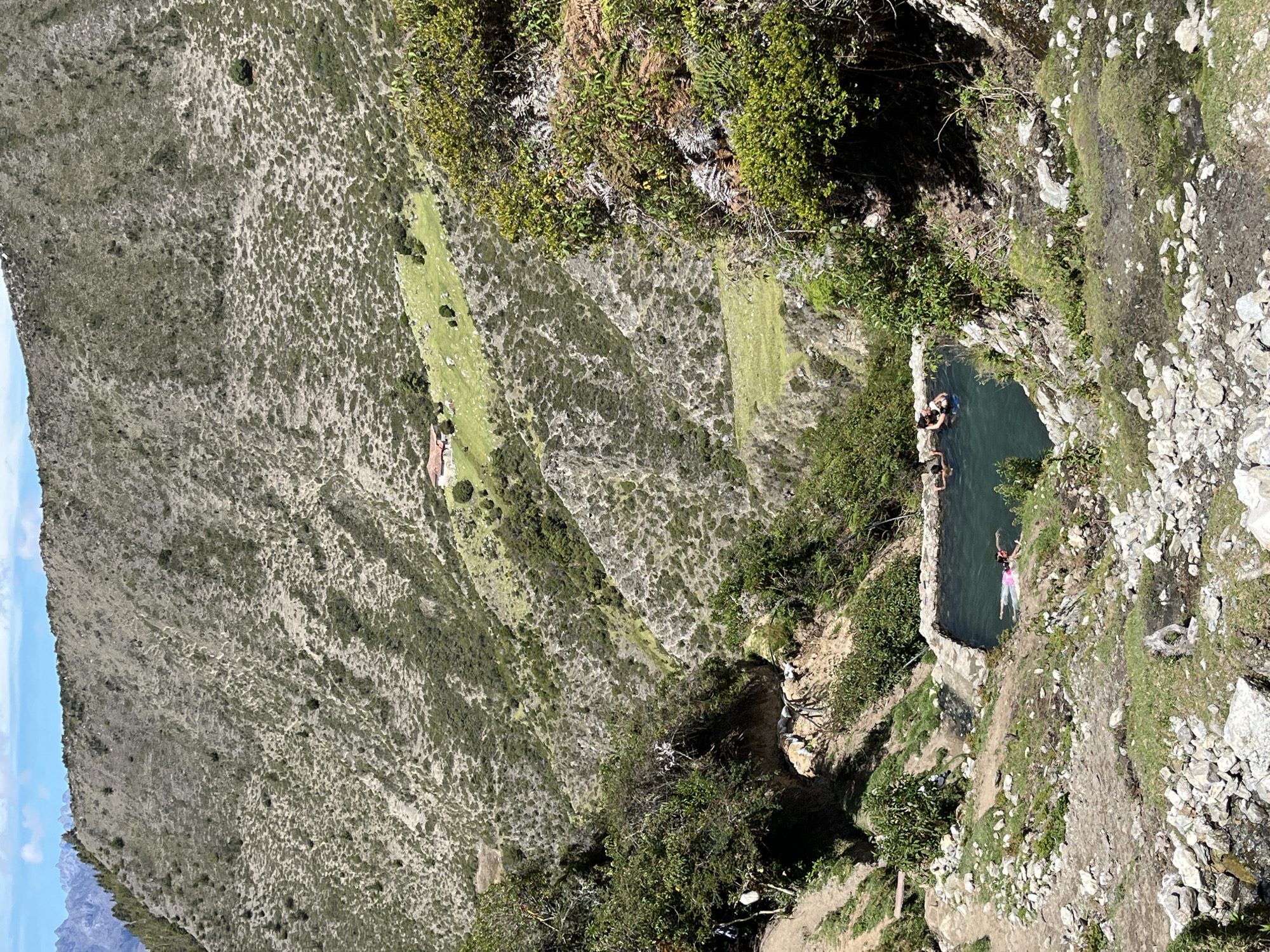 The image shows a natural hot spring pool in a mountainous, rural area. The pool, constructed with stone borders, is filled with dark blue water and has a few people swimming in it. One person in the foreground is swimming on their back wearing a pink swimsuit. The surrounding environment is rugged and natural, with steep, vegetated slopes and rocky terrain. In the distance, a small house with a red roof is visible on a grassy hillside. The sky above is blue with scattered white clouds. The location is identified as a hot spring in Mucurubá, Venezuela.