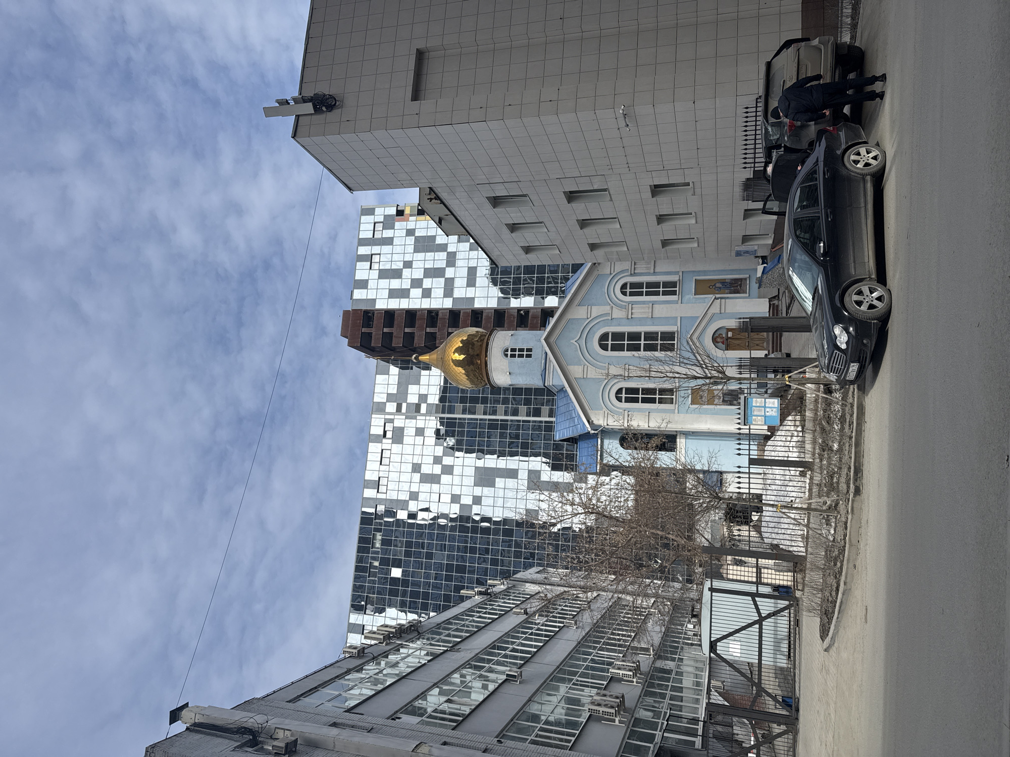 The image depicts a street scene in Yekaterinburg, Russia. A light blue orthodox church with a golden dome stands adjacent to modern buildings, including one with a checkered facade. A black Mercedes-Benz is parked on the street, with two individuals near the vehicle. Fences surround the church grounds. The sky is partially cloudy.