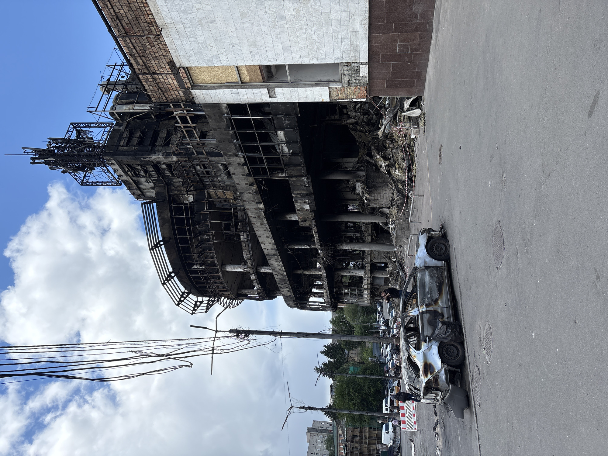 The image depicts the charred remains of the Retroville shopping mall in Kyiv, Ukraine, following a bombing attack. The building's facade is extensively damaged, with blackened skeletal structures exposed. A burnt-out car sits on the roadside in front of the building. Electrical wires hang haphazardly from damaged poles. The sky above is partly cloudy.