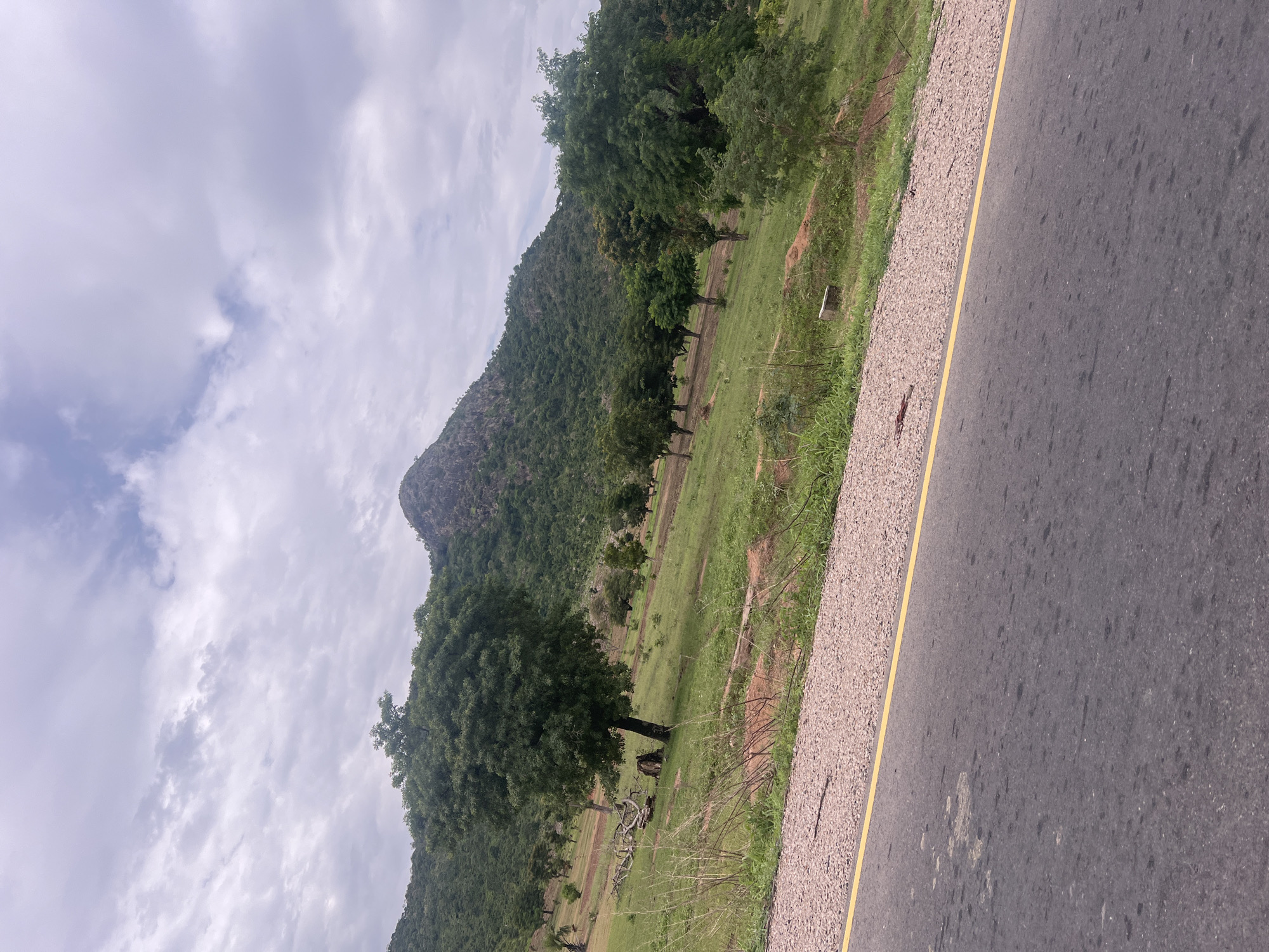 The media file captures a scenic view of a mountainous landscape in Minda, Nigeria. The foreground features a paved road with a distinct yellow line marking its edge, bordered by a gravel shoulder. Beyond the road, a lush green grassy field stretches out, dotted with several trees. A prominent hill or mountain, covered in dense vegetation, dominates the mid-ground and background. The sky above is cloudy, suggesting a possibly overcast or approaching weather event. The scene appears to be a typical rural or natural setting, likely during daylight hours given the ambient light. There is no discernible activity or event taking place, and no individuals or specific objects are highlighted beyond the natural elements and the road. The overall impression is one of natural beauty and tranquility.