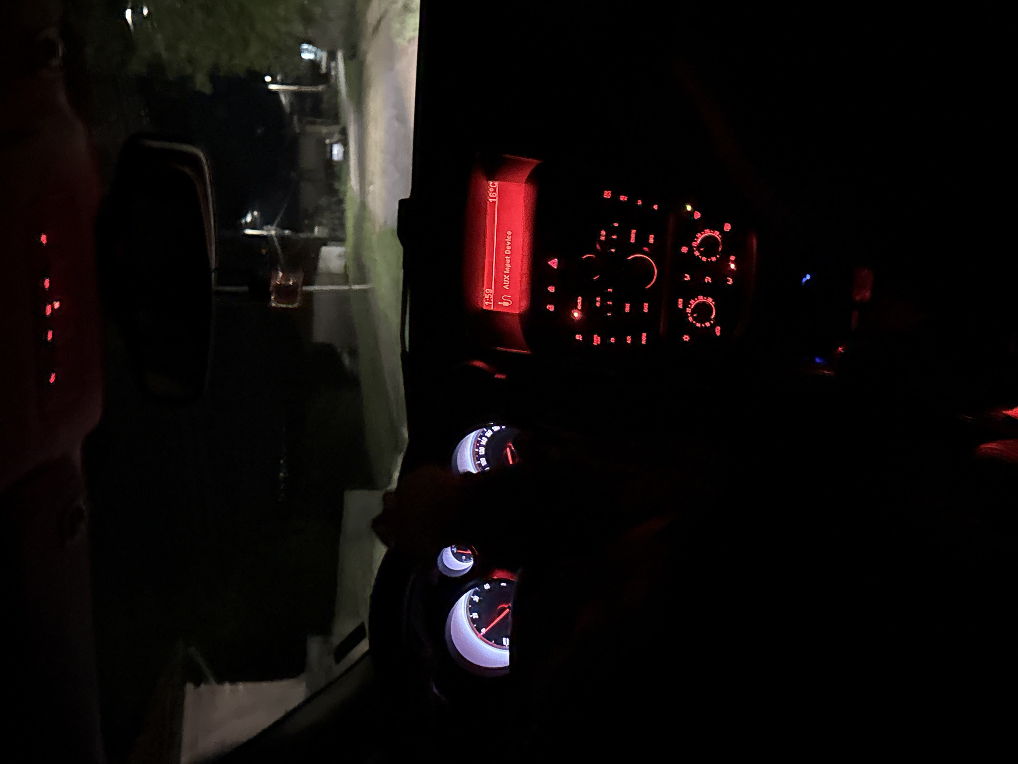 The image shows the interior of a car at night, with the dashboard and instrument panel illuminated. The radio display shows "AUX Input Device" and a temperature of 16°C. The speedometer and other gauges are visible but not clearly readable. Outside the car window, a dimly lit street and buildings can be seen. The location is not evident from the image.