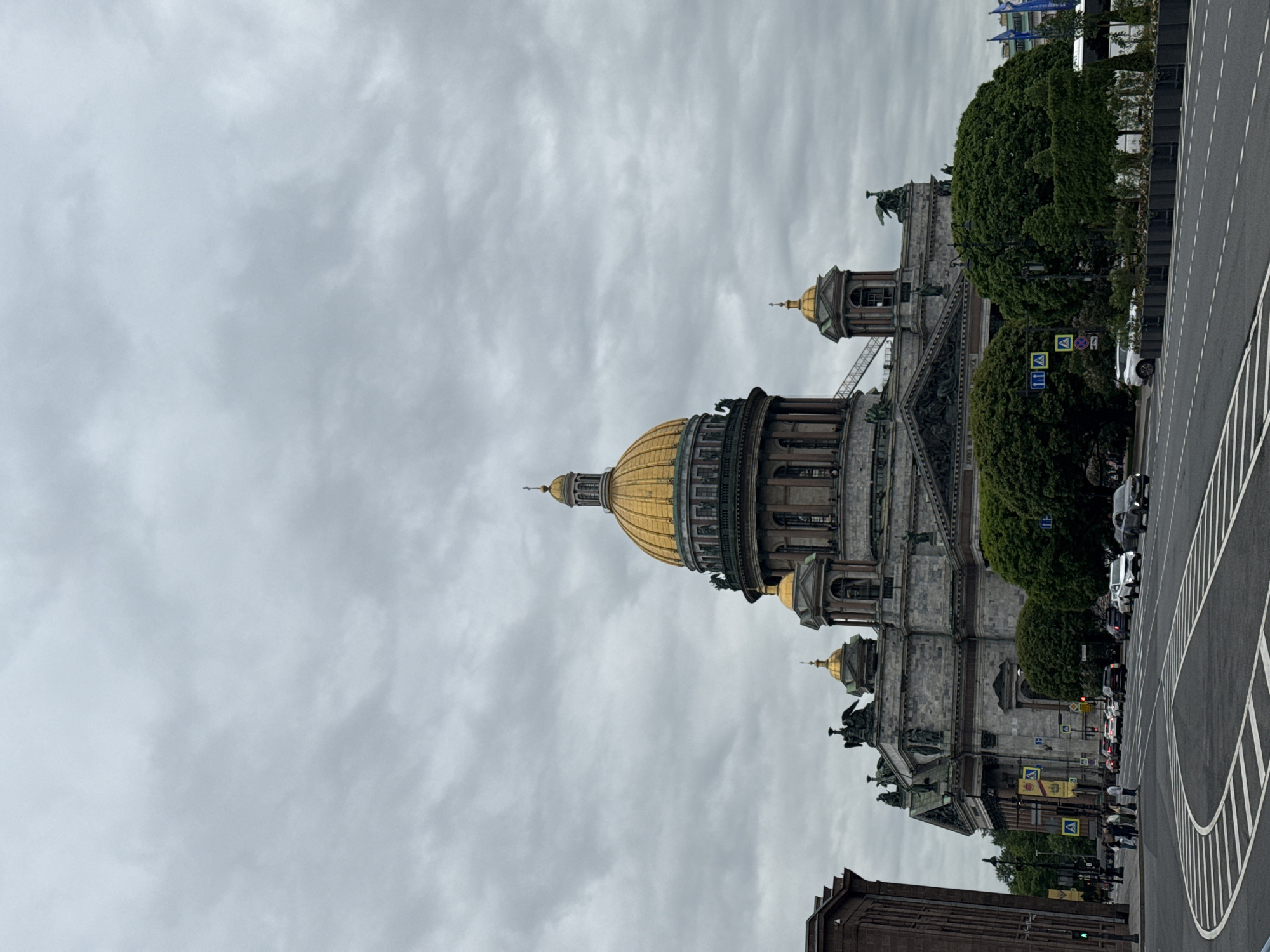 The image depicts St. Isaac's Cathedral in Saint Petersburg, Russia, under a cloudy sky. The cathedral's golden dome and gray facade are prominent features. The street is visible with traffic, pedestrian markings, and signage. The presence of trees partially obscures the building's facade, and some statues are visible atop the building.