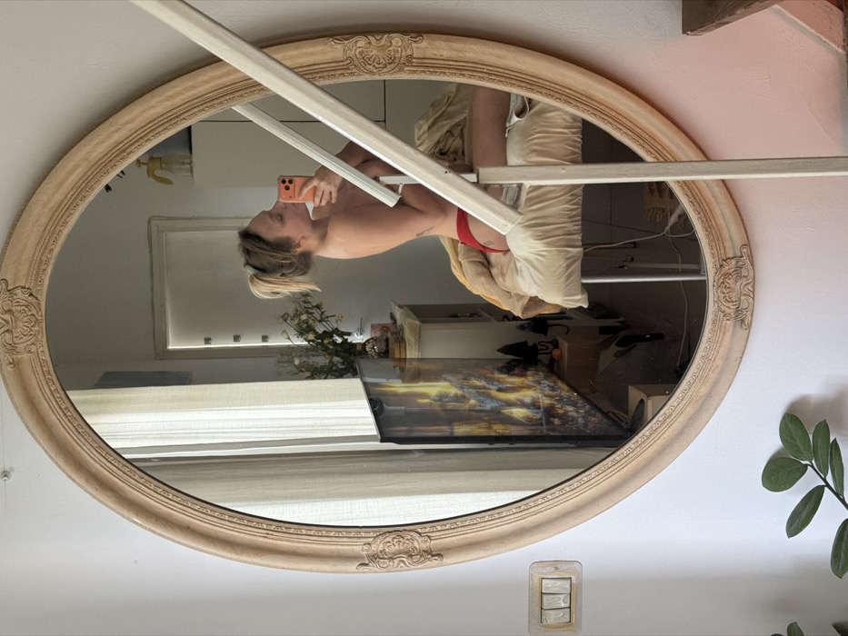 The image captures an interior scene, confirmed to be in Tel-Aviv, Israel, showing a large oval mirror with an ornate, light-colored frame. The mirror is mounted on a white wall, revealing a discolored triple light switch to its immediate left and a green leafy plant at the bottom left.
Reflected in the mirror is a female individual with light-colored hair pulled back in a ponytail. She is bare-chested and wearing red briefs, with a small tattoo visible on her left hip/pelvic area. She holds a smartphone with an orange case to her face, actively taking a mirror selfie. Her body is positioned on a light-colored, padded surface, covered with a beige blanket or sheet.
The mirror's reflection also displays parts of the room: a television screen to the left showing an image of fire or a vibrant abstract pattern, a light-colored cabinet or dresser with a vase of dried flowers and other small objects, and a doorway in the background. A window with light-colored curtains is visible to the left within the reflection, suggesting natural light. A yellow spray bottle is visible on a shelf in the background. A light-colored staircase railing extends diagonally across the lower right portion of the image, partially obscuring the reflection of the individual and the padded surface. A dark wooden surface is visible at the very bottom right, adjacent to the staircase.