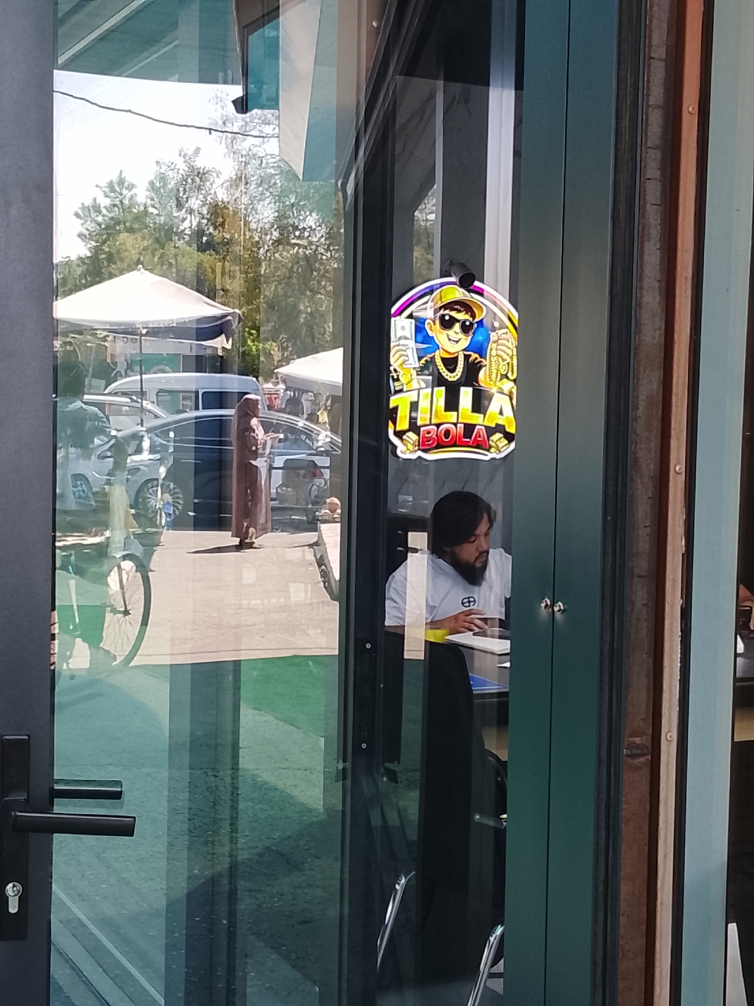 This image captures a street scene in Fergana City, Uzbekistan, viewed through a glass door. The foreground features a dark grey door handle and lock on the left. Beyond the glass, a brightly colored sign depicting a cartoon character wearing sunglasses, a gold chain, and holding money is prominently displayed. Below this sign, a man with dark hair and a beard is seated at a desk, looking down at a notebook or papers. He is wearing a white t-shirt with a circular logo. Reflections on the glass show trees and market stalls, including white tents and parked vehicles. A person in a long brown garment is visible standing among the stalls. A bicycle is also reflected in the glass, partially visible on the left. The environment appears to be an outdoor market or street area.