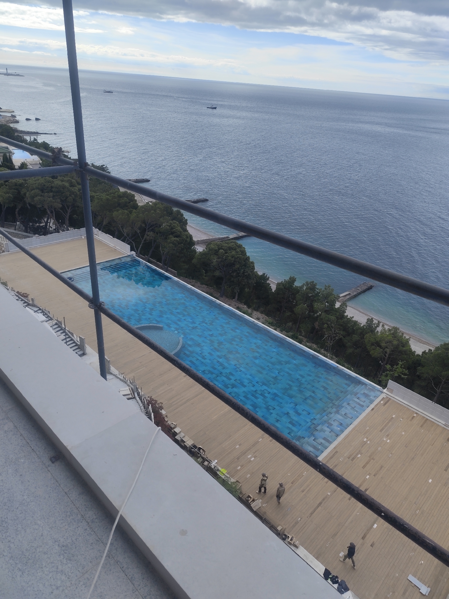 The image shows a partially constructed outdoor swimming pool located near the coast in an unknown city. The pool is rectangular in shape with blue tile and a circular section at one end. A wooden deck surrounds the pool. Three individuals are visible on the deck; two are standing close together, and the third is walking. Scaffolding is present in the foreground, partially obscuring the view. In the background is the ocean with visible coastline and scattered trees along the shore. The sky is overcast. The exact location within the city and country is not discernible.