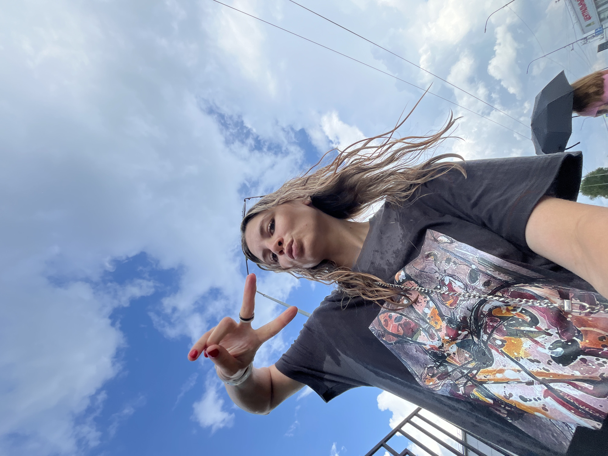 A young woman with long, wet, blonde hair is taking a selfie in Chaikovsky, Russia. She is wearing a dark t-shirt with a colorful graphic print and a silver chain necklace. Her right hand is raised, with her index and middle fingers extended in a peace sign. She is puckering her lips. The background shows a cloudy blue sky and some power lines. In the distance, another person is walking under a black umbrella. A metal railing is visible on the left side of the frame.
