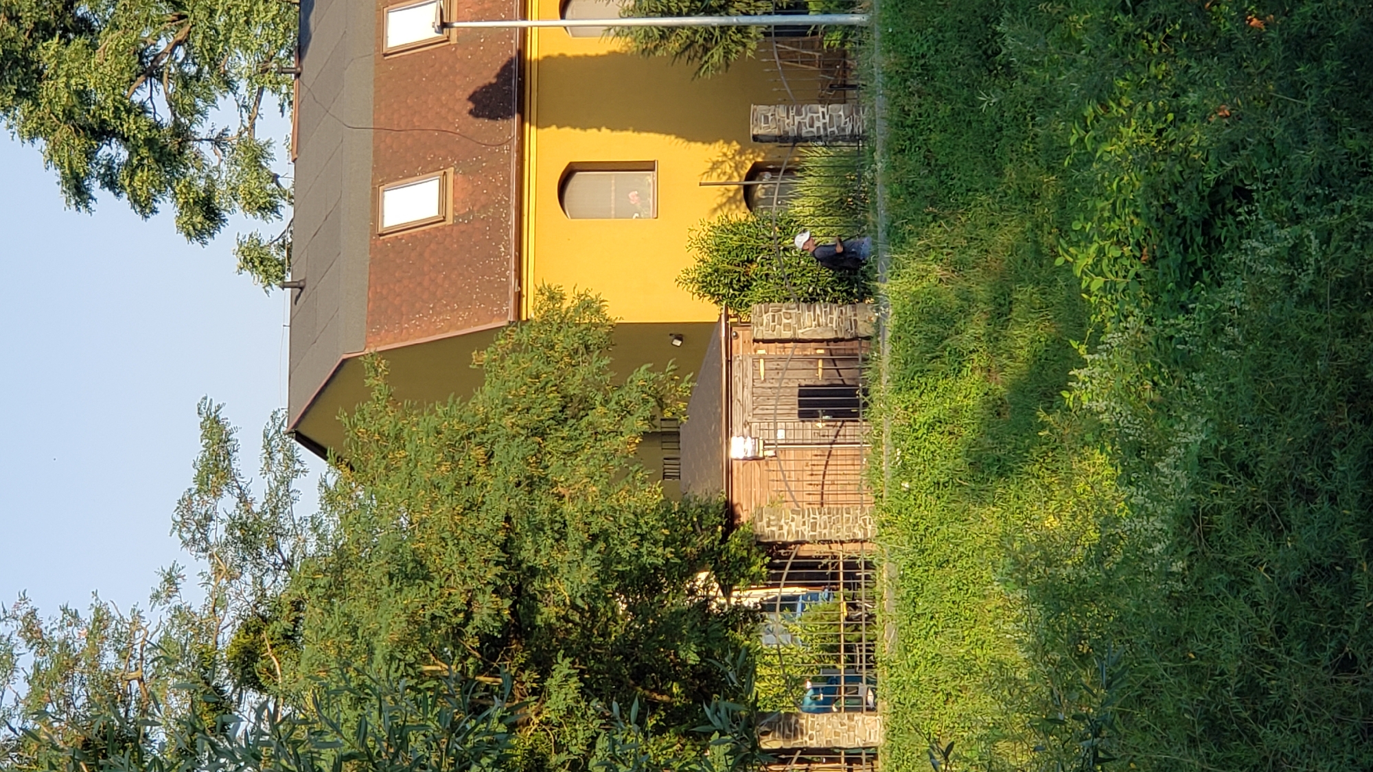 This is a photograph of a yellow building with a brown, tiled roof and several dormer windows, located in Prešov, Slovakia. In front of the building is a small, wooden shed with a dark, sloped roof, constructed with stone pillars at its corners. A metal fence encloses the area around the shed. A person wearing a white cap and a dark backpack is walking along a path near the fence. The area is surrounded by lush green grass and trees, with sunlight casting shadows on the ground. The sky is clear and blue.