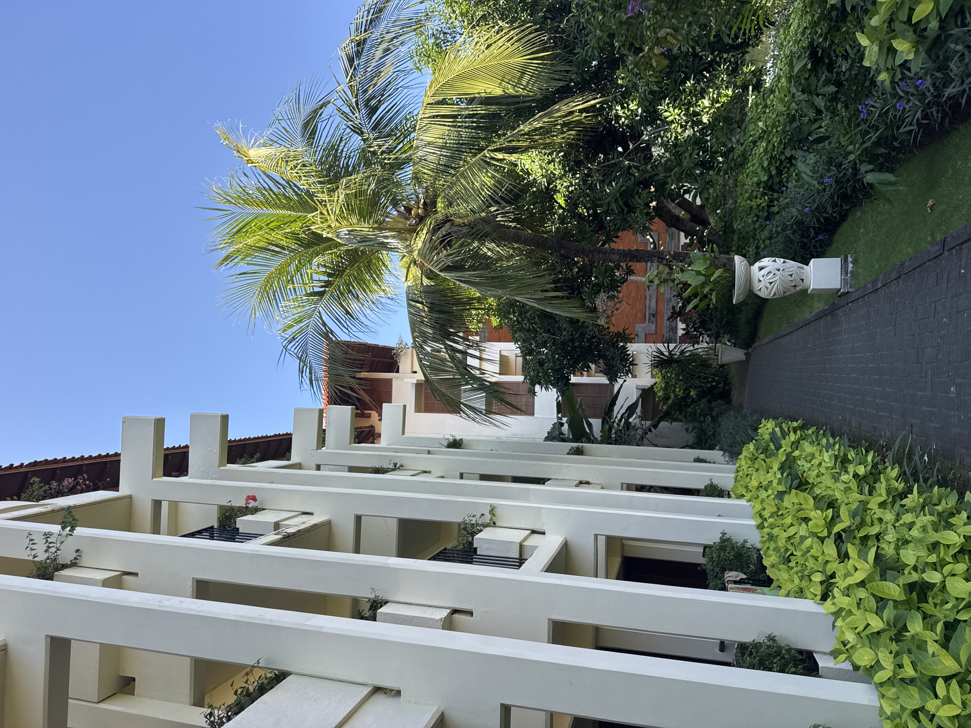 In Kuta Selatan, Indonesia, a white, modern building with a multi-level facade features open balconies and vertical support beams, overlooking a paved walkway. Lush green foliage lines the walkway, with a palm tree and other tropical plants visible in the background. A white, ornate vase with a plant sits on a pedestal beside the path. The sky is clear and blue.