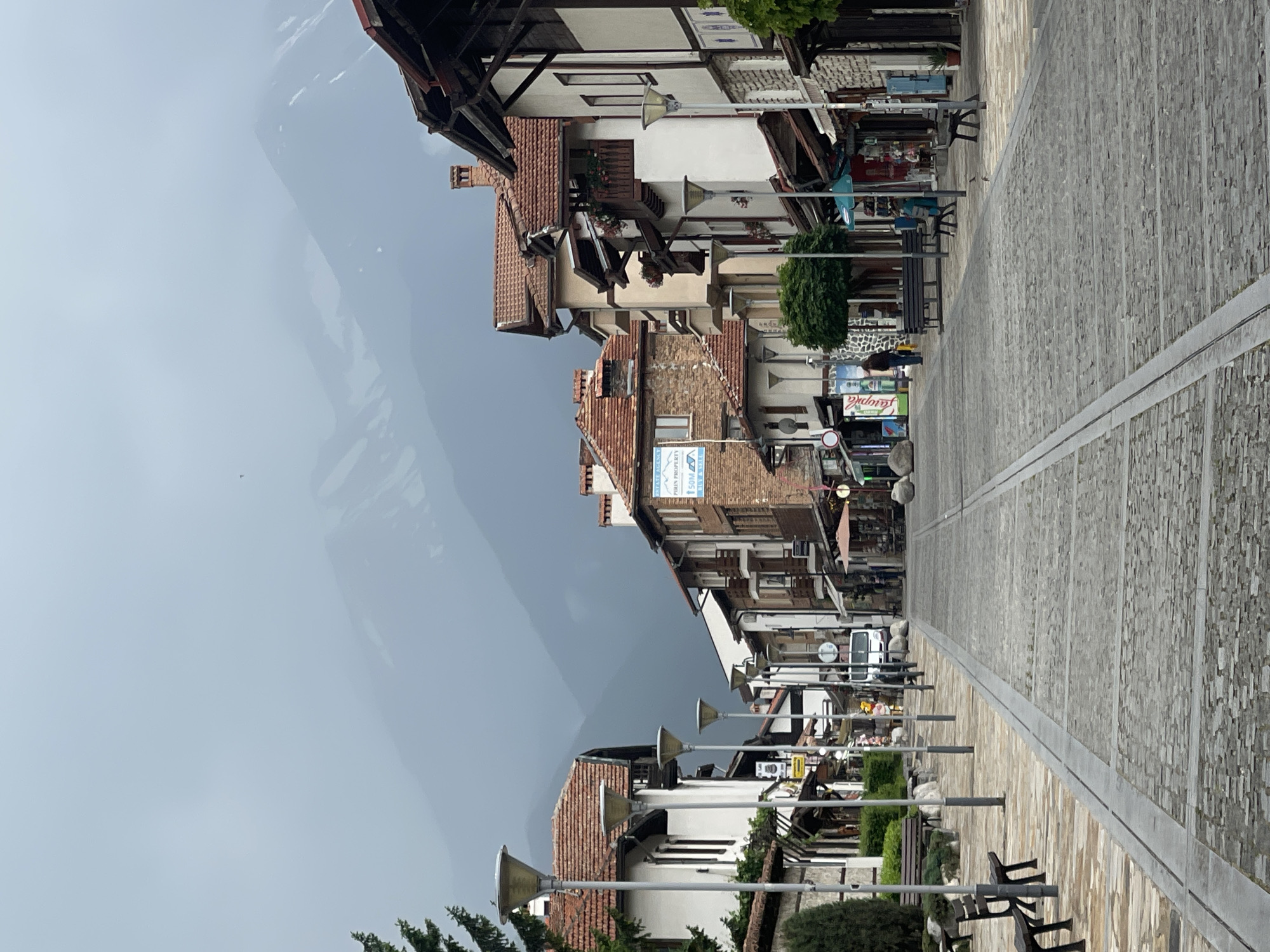 The image depicts a European-style cobblestone street lined with traditional buildings and a hazy mountain range in the background. The street is paved with grey cobblestones and has two parallel metallic tracks running down the center. On the left side of the street, there are white buildings with red tile roofs, lampposts, and benches. A tall evergreen tree is visible on the far left. On the right side, buildings with red tile roofs, wooden balconies, and shops are present. Some people are walking on the street, and a white van is parked in the distance. The sky is overcast and a light mist obscures the mountain peaks. The overall impression is one of a quiet, possibly historic, town or village.