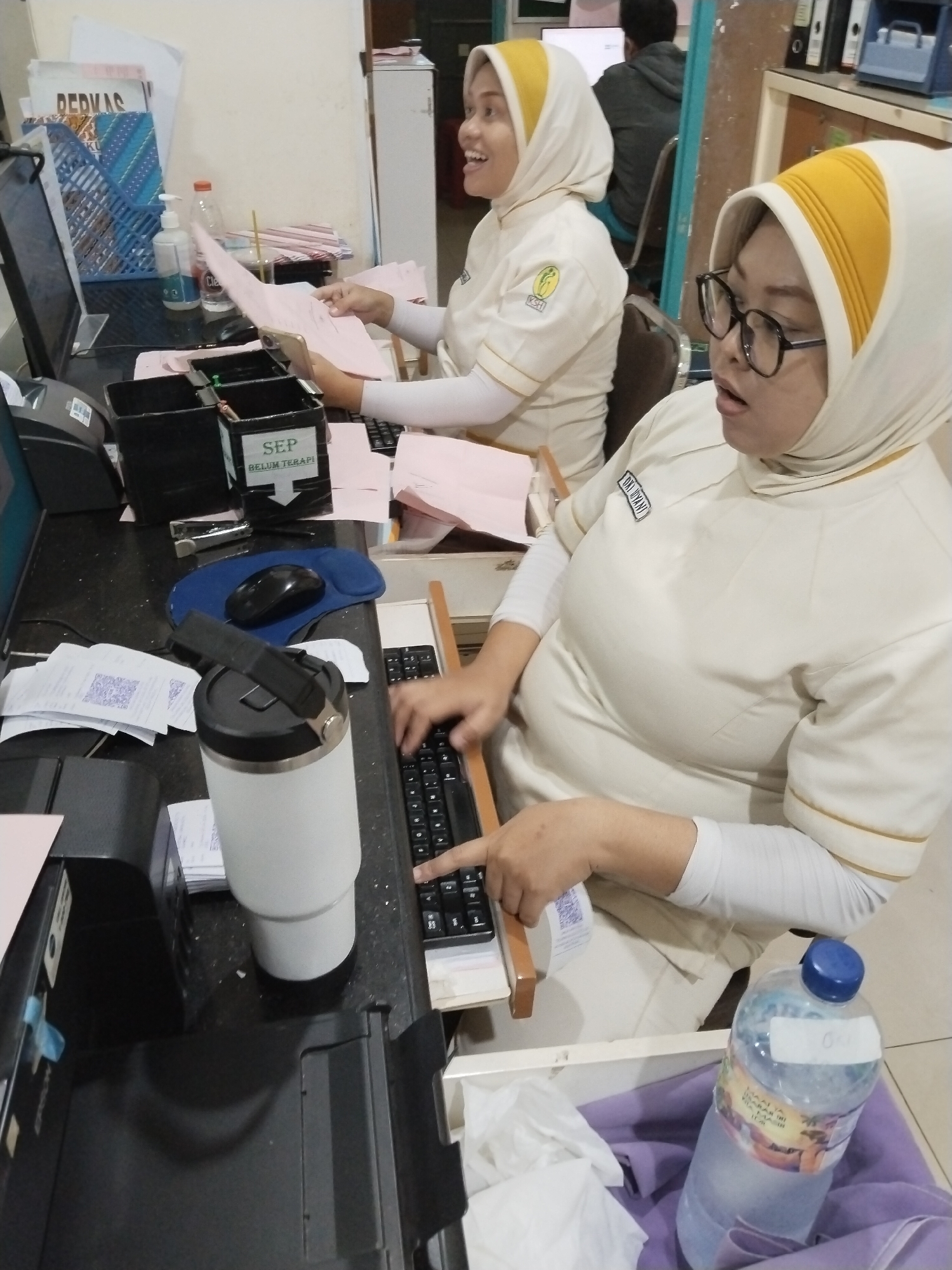In Margorejo, Indonesia, two women wearing yellow and white uniforms with headscarves are seated at desks in what appears to be an office environment. The woman on the right, wearing glasses, is actively typing on a keyboard. In front of her is a large white insulated tumbler. To her right, a partially filled plastic water bottle with a blue cap sits on a purple cloth. Various papers, including receipts with QR codes, are scattered on the desks. A black stapler and a blue mousepad with a black computer mouse are also visible. A black printer is situated to the left of the frame. The woman on the left is holding pink papers and looking to her right. Behind them, a man wearing a grey hoodie is visible in the background, seated in front of a computer screen. The environment is cluttered with office supplies and equipment, suggesting a busy workspace.