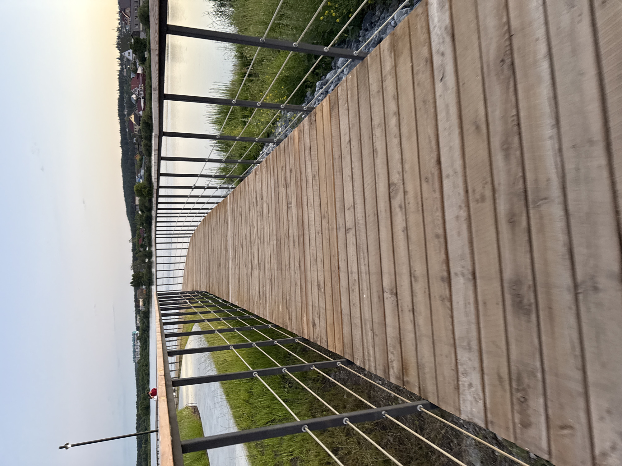 This is a photograph of a wooden boardwalk that curves along the edge of a body of water. The boardwalk is made of light-colored wooden planks laid vertically. A black metal railing with thin, taut wires runs along the edge of the boardwalk, separating it from the water. To the left of the boardwalk, there is a grassy area with some rocks and yellow wildflowers. Beyond the grass, a calm body of water reflects the sky. In the distance, across the water, there is a treeline and a town with buildings. One of the buildings appears to be a multi-story apartment complex. The sky is a pale, hazy yellow, suggesting it is either sunrise or sunset. The overall atmosphere is peaceful and serene. In the foreground, a person wearing a red shirt can be seen sitting on the edge of the boardwalk, looking out at the water. The scene appears to be a public park or recreational area. There is no visible text in the image. The photograph is taken from a slightly elevated perspective, looking down the length of the boardwalk. The lighting suggests it is a calm, overcast day or early morning/late evening.