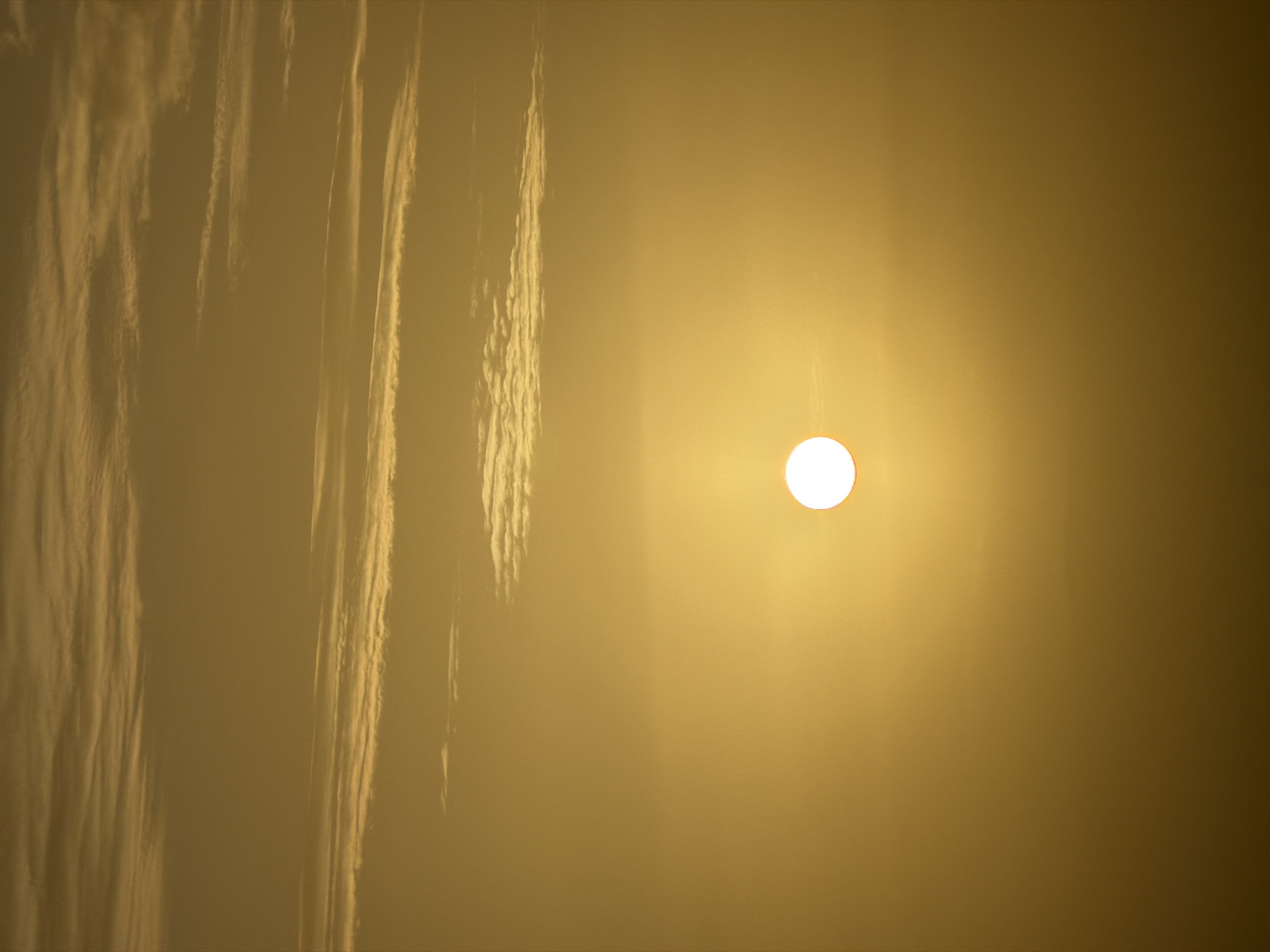A close-up shot shows a hazy, golden sky with streaks of clouds on the left. The bright sun is positioned towards the right, appearing as a white circle with a slight reddish outline.