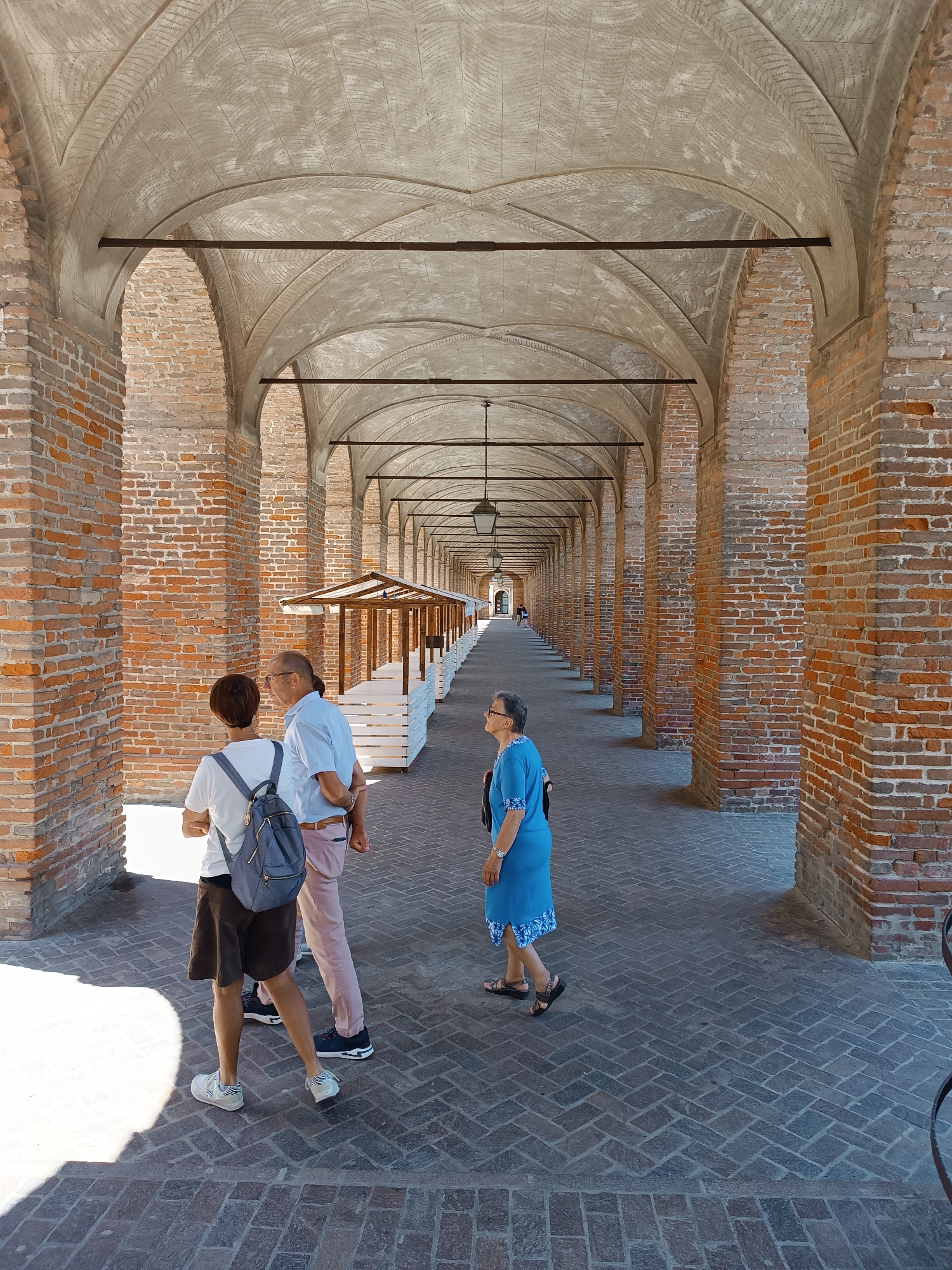 The image shows an open arcade with a vaulted ceiling and brick pillars, likely a historic building in Mezzana Sant'Antonio, Italy. Three people are present: two, a person with short dark hair in a white shirt and backpack and a person with a light blue shirt, are standing together facing away from the viewer. An older person in a blue dress is walking a few steps ahead, away from the viewer. A row of market stalls sits parallel to the people on the left. The corridor extends far into the distance, with the image exhibiting a strong sense of depth and vanishing point. The floor is paved with bricks, and the overhead lighting consists of hanging lanterns. The scene is bathed in sunlight.