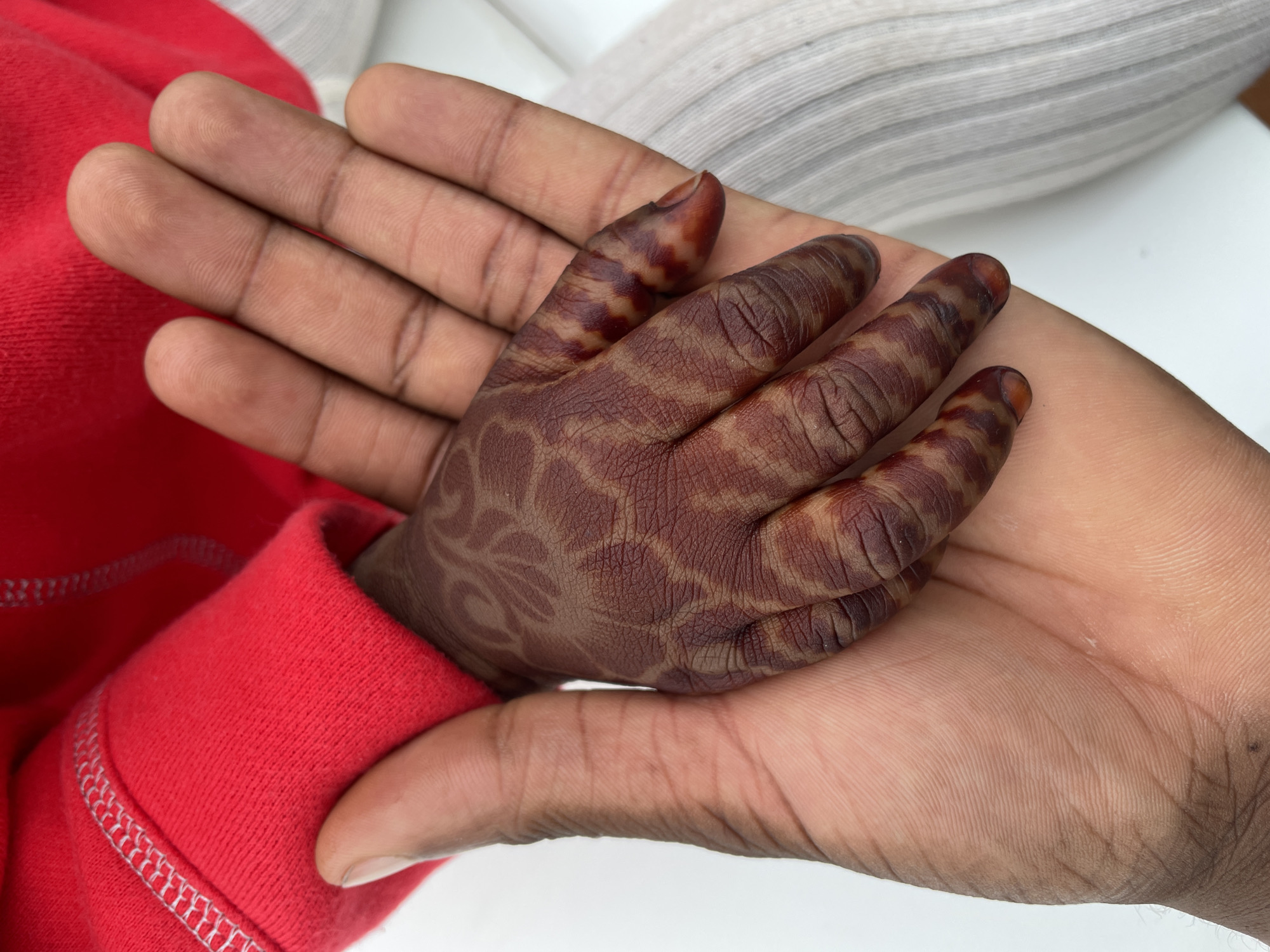 A fair-skinned adult hand holds a child's smaller hand that is patterned with a henna-like dye. The child's hand is clad in a red garment with a white-stitched cuff. The background includes a white surface and a striped white garment. No landmarks indicate a location in Barakin Akawo, Nigeria.