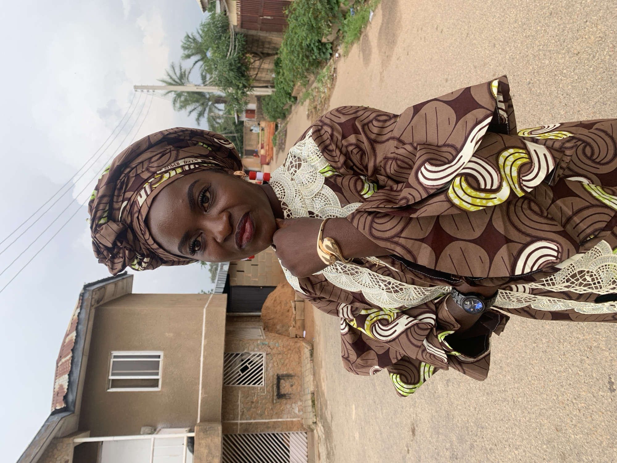 A medium shot captures a Black woman in traditional African attire standing on an asphalt road in front of a two-story building. The woman wears a brown headwrap and a brown, white, and green patterned dress with flared sleeves. She has on red and white earrings and a gold bracelet, and a black wristwatch. Her left hand is resting on her chin, and her right hand is on her chest. The building behind her has a brown stucco exterior and a rusty corrugated metal roof. The woman is positioned in the foreground, facing slightly to her right. The background shows a residential street with vegetation and more buildings. The sky is overcast.