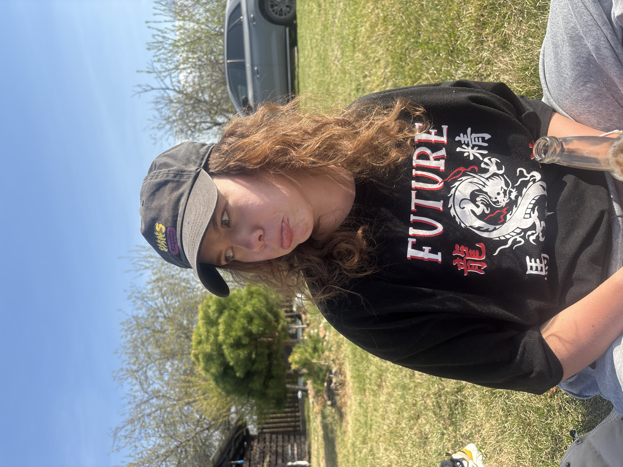 The photograph shows a person, likely female, sitting on a grassy surface outdoors. They are wearing a black cap with "VANS" visible and a black long-sleeved shirt with a dragon and the word "FUTURE" printed on it. Their hair is curly and light brown. A bottle is visible near their right side. The background features a clear blue sky, trees with sparse foliage, a gray car, and a small wooden structure. The location may be Emel'yanovo, Russia, based on provided information, though the photo alone does not provide direct evidence to confirm the location.