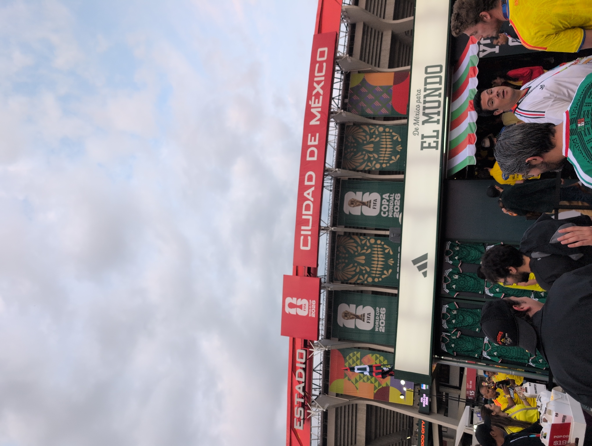 The image shows the exterior of a stadium in Mexico City, Mexico, with banners promoting the FIFA World Cup 2026. The stadium has a prominent red facade with large white lettering that reads "ESTADIO CIUDAD DE MÉXICO". Below the stadium's main structure, a retail stall with an Adidas logo is visible, featuring green soccer jerseys for sale. Several people are gathered around the stall, some wearing soccer jerseys themselves. The atmosphere appears to be festive, suggesting an event related to the upcoming World Cup.