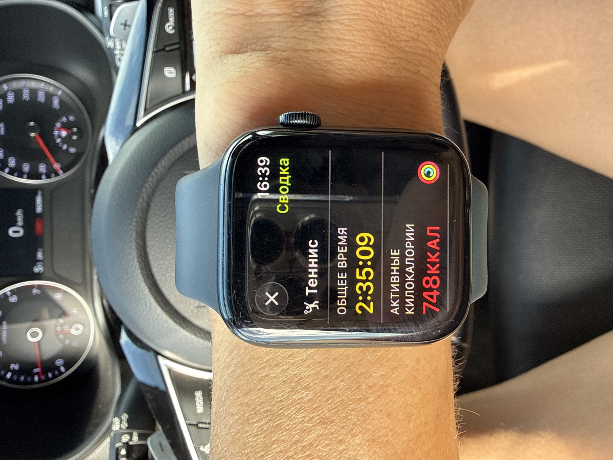 The image shows a close-up of a person's wrist wearing an Apple Watch, which is displaying a summary of a tennis workout. The workout summary includes the total time spent playing tennis, which is 2 hours and 35 minutes and 9 seconds, and the active calories burned, which is 748 kcal. The watch face also shows the current time as 16:39. In the background, a steering wheel and dashboard of a car are visible. The car is likely parked in a city, but there is no specific information to determine if it is in Chaikovsky, Russia. The lighting suggests it is daytime. The overall scene is of a person checking their fitness data after a workout, possibly while in their car.