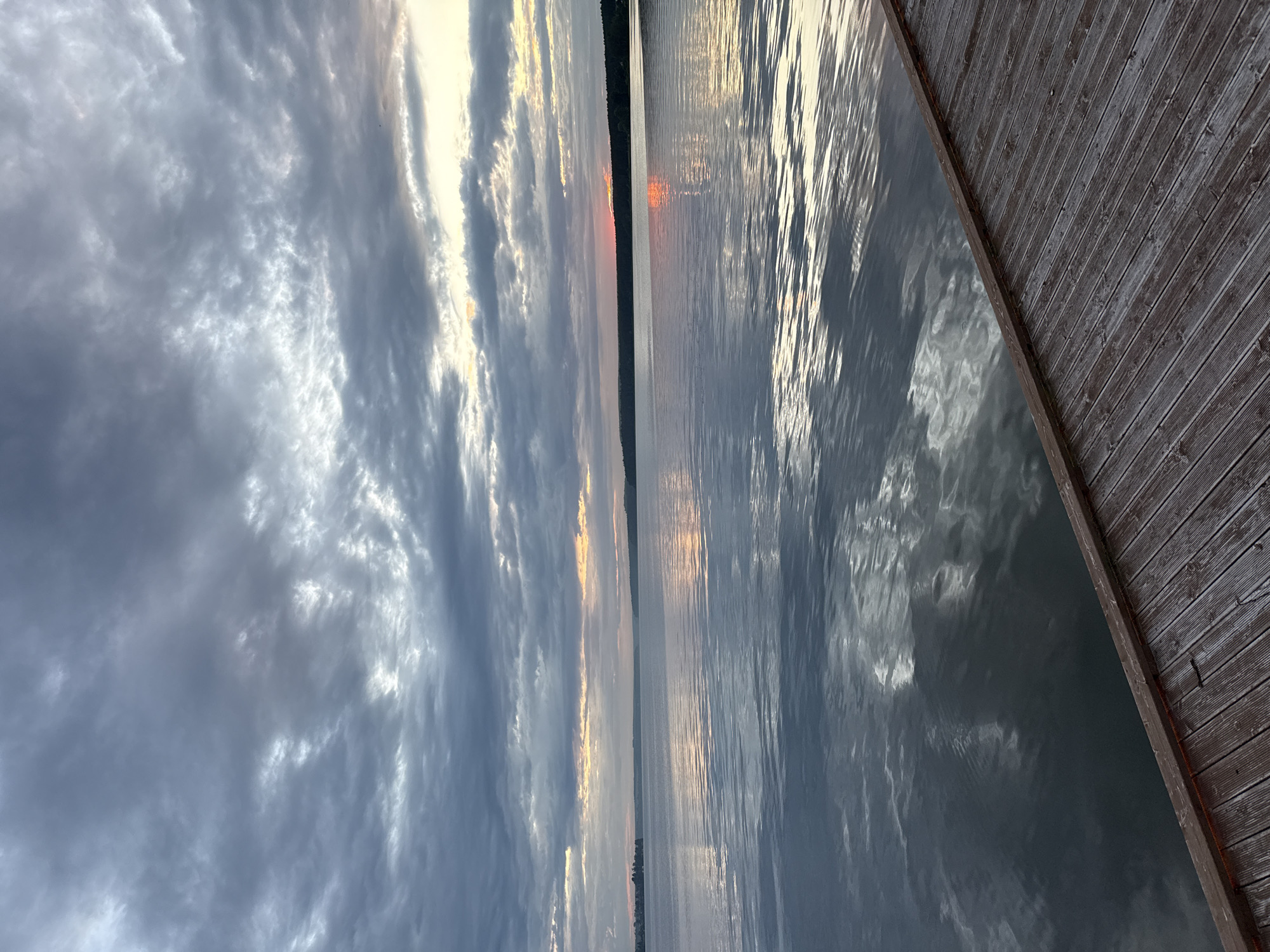 The image depicts a serene lake scene, likely at dusk or dawn, given the soft, diffused light and the reflections on the water. The sky is filled with dramatic, heavy clouds that are tinged with orange and yellow hues, suggesting the presence of the sun just below the horizon. The water's surface is calm, with gentle ripples that catch and reflect the light and the colors of the sky. In the foreground, a wooden dock or pier extends into the water, its weathered planks adding a rustic texture to the scene. The distant shoreline is lined with dark trees, creating a silhouette against the brighter sky. The overall impression is one of tranquility and natural beauty. This scene could be located on Lake Votkinsk, which is a significant body of water in the city of Votkinsk, Russia.