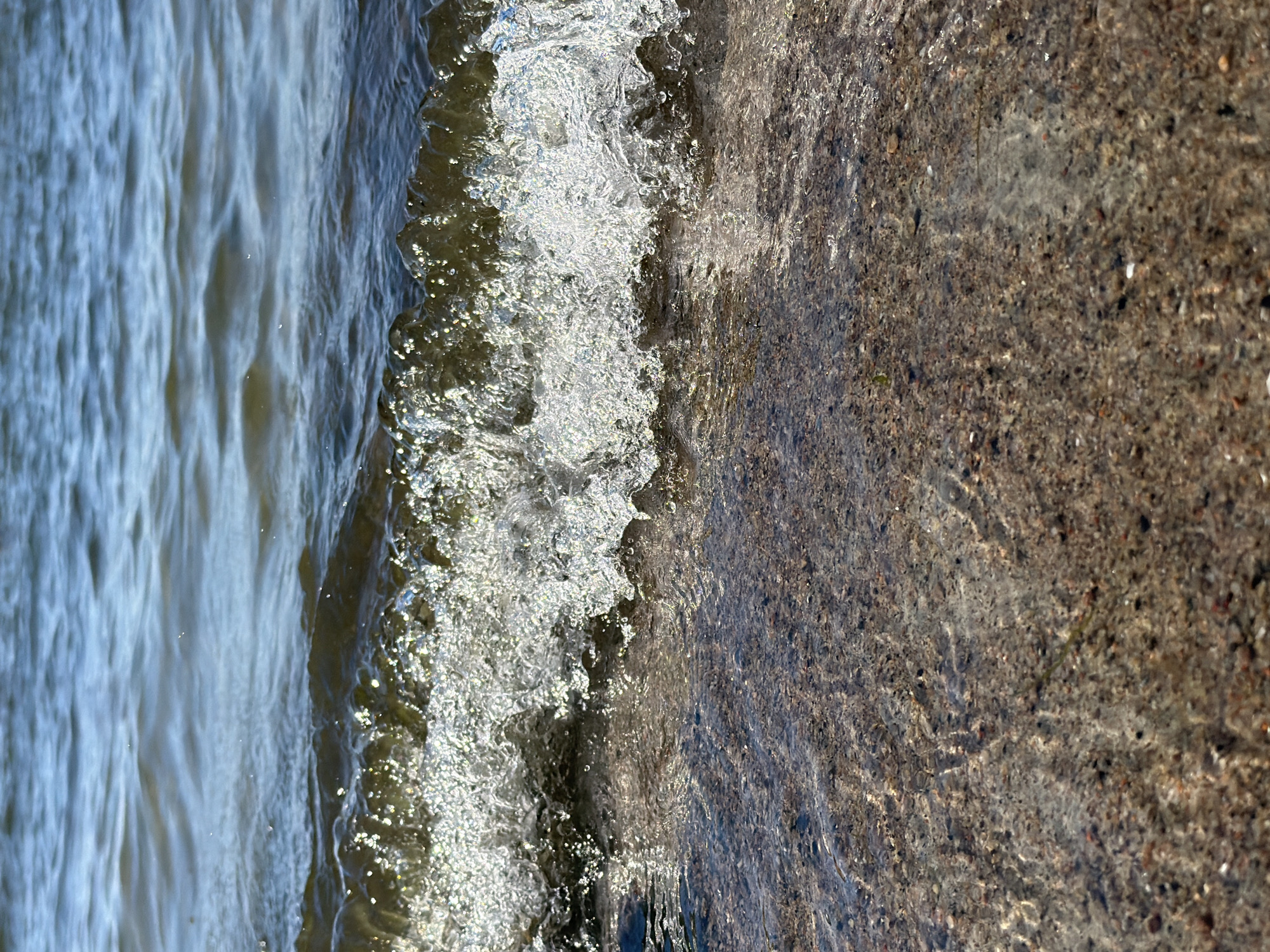 The image shows a close-up view of a wave gently breaking on a pebble beach. The water is clear, allowing the viewer to see the various sized pebbles and small stones beneath the surface. Sunlight reflects off the water's surface, creating a shimmering effect. The background shows the choppy surface of a larger body of water, with ripples and waves stretching towards the horizon. The scene appears to be a natural outdoor environment, likely a shoreline or beach. Without further context or identifying markers, it is not possible to determine if this specific location is in the city of Tallinn, Estonia.
