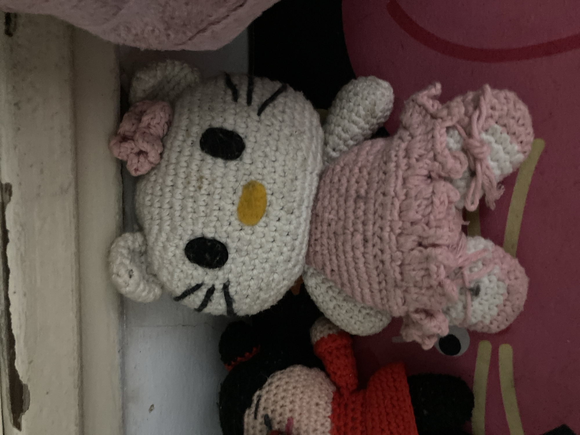 The image displays a close-up, rotated view of several crocheted plush toys in an indoor setting. The most prominent subject is a white, Hello Kitty-like doll with distinct black oval eyes, a yellow oval nose, and black embroidered whiskers. A small pink crocheted bow is attached to its left ear. The doll's body is predominantly pink, with white accents on its feet or lower legs, suggesting shoes.
Partially visible below the Hello Kitty doll is another crocheted toy, resembling Minnie Mouse. This doll has a black body and head, with a light pink face and a red element that could be a hat or part of its attire.
The dolls are positioned on a dark pink surface, possibly a rug or mat. To the left of the toys, a light-colored, textured background, resembling a worn wall or furniture, is visible. A soft, light pink crocheted or knitted fabric is also present in the upper left corner of the frame. The intricate texture of the yarn and the crocheted stitches are clearly discernable on all the toys, highlighting their handmade nature. There is no specific activity taking place, and no text is visible in the image.