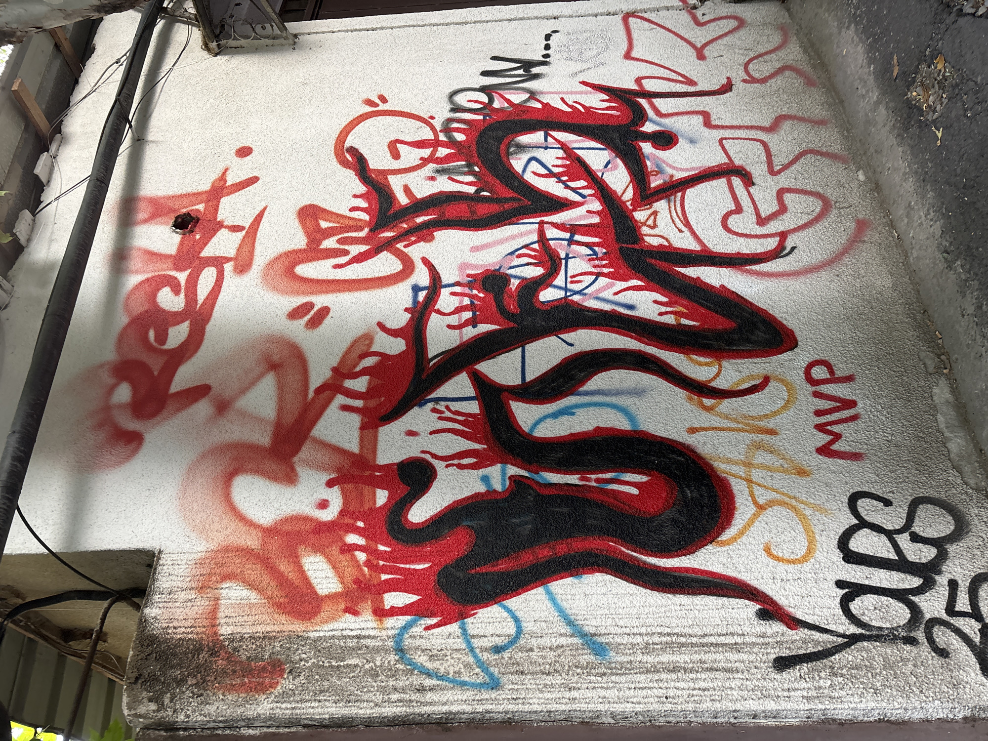 This media file captures a close-up, high-angle shot of graffiti on a textured white wall. The artwork primarily features abstract, flowing shapes in vibrant red and black, with hints of other colors like blue and yellow spray-painted in the background. The style is dynamic and energetic, with sharp lines and curved forms suggesting movement.
The graffiti appears to be layered, with older, faded red spray paint visible beneath the more prominent black and red figures. One section of the graffiti, on the right side of the image, contains legible text. These include "MVP" in red, "Jdbs" in black cursive, and a partial word that might be "AS." There are also some smaller, less decipherable tags.
The wall itself shows signs of wear and tear. A concrete ledge or beam runs along the bottom of the image, and above it, the wall is rough and speckled. The top of the image shows the edge of a roof or overhang with some exposed metal and electrical wiring. The lighting suggests it might be daytime, but the specific time and weather conditions are not discernible. The overall impression is of an urban or industrial setting where graffiti is prevalent. No people are visible in the frame.