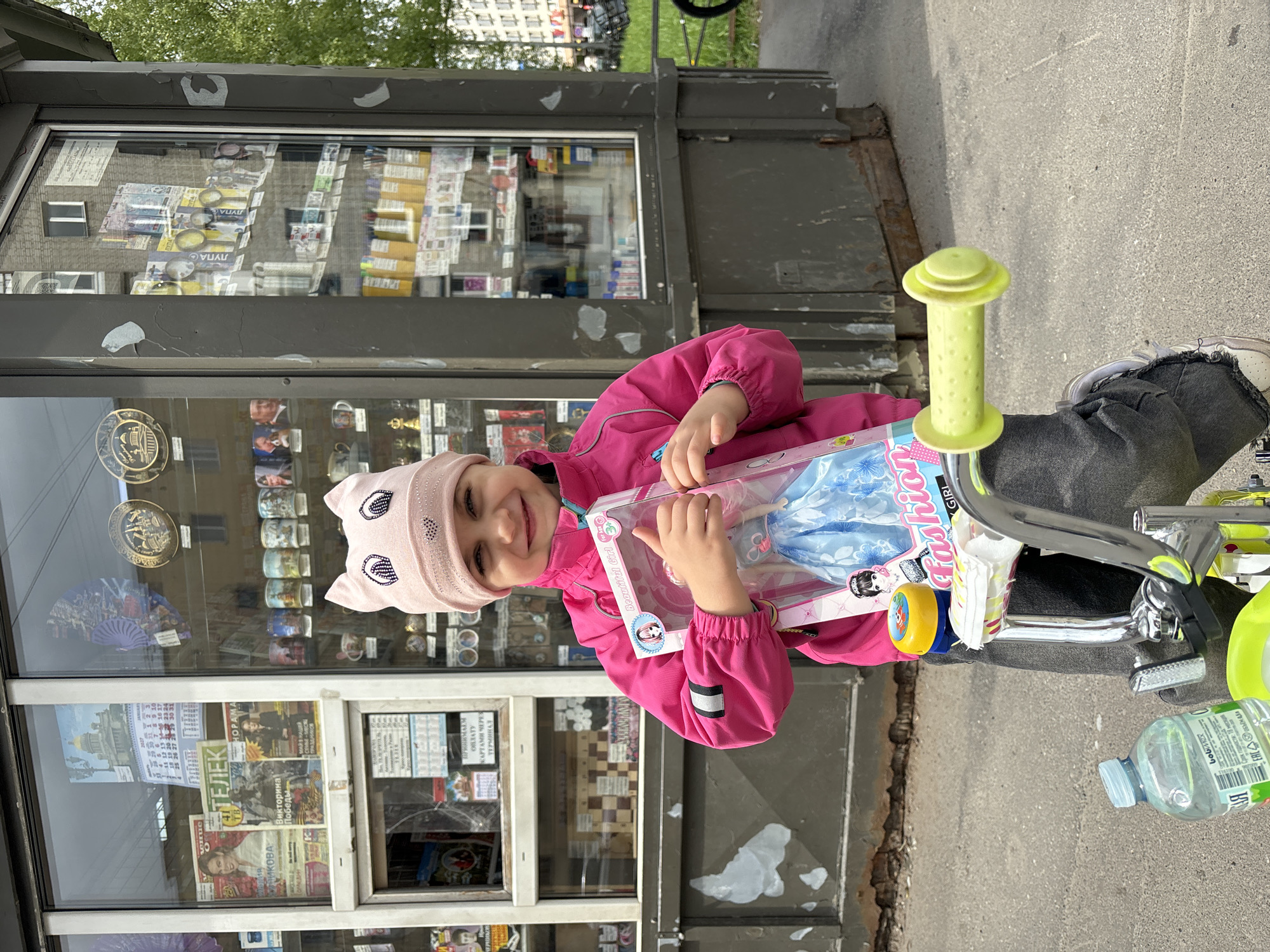 A young girl in a pink jacket and hat stands next to a kiosk on a street. She is holding a doll in a box. A bicycle is visible to the right of the girl. A water bottle is on the ground next to the bike. The kiosk displays various items including newspapers, calendars, and souvenirs. The photograph appears to be taken outdoors in Saint Petersburg, Russia, as indicated by the writing on the kiosk displays.