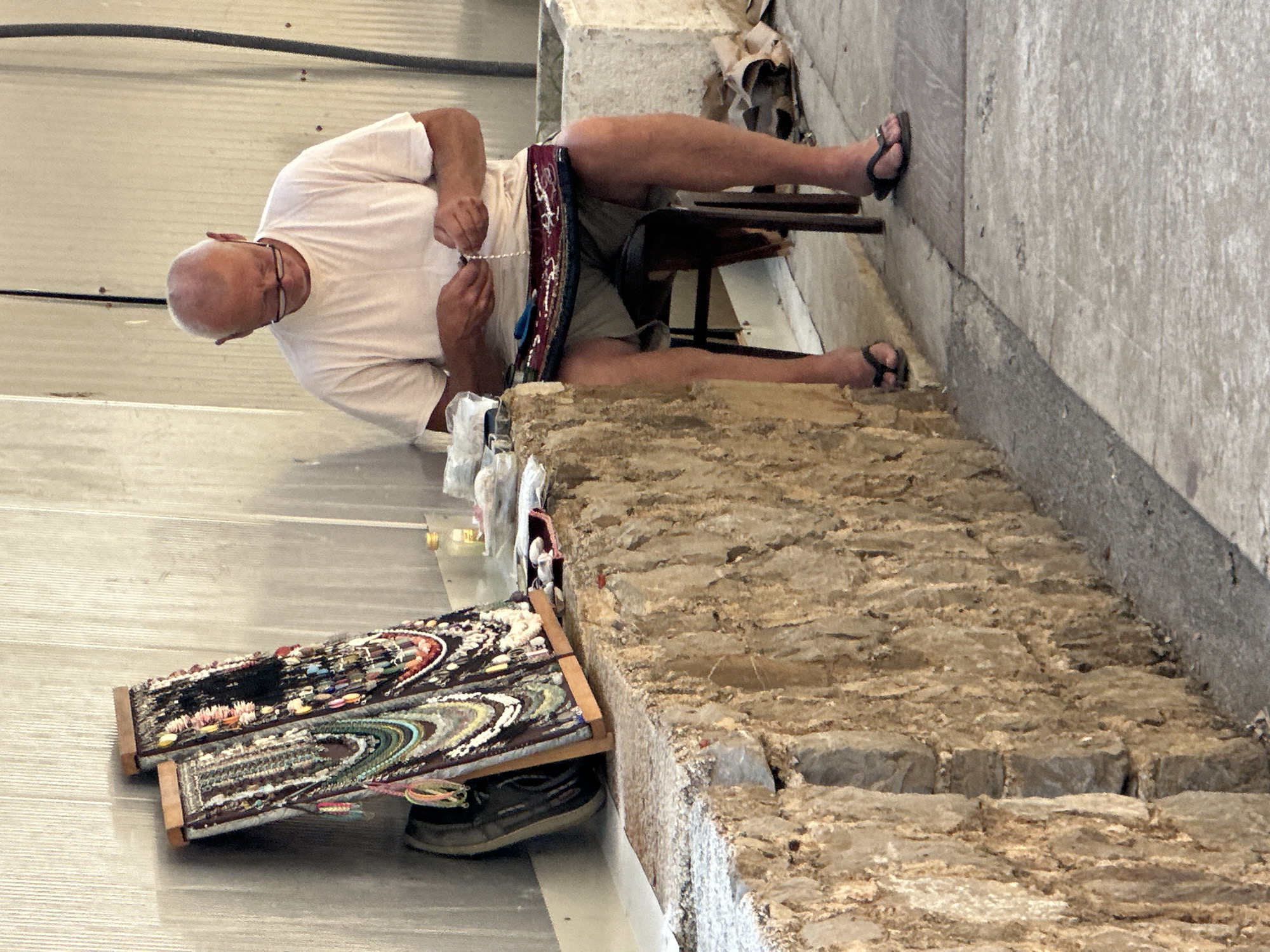 A middle-aged, bald Caucasian man wearing a white t-shirt and khaki shorts sits on a dark stool, facing away from the camera. He is wearing glasses and appears to be stringing beads, holding a strand of pearls in his hands. A piece of fabric with intricate beadwork is draped over his lap. To his left, a wall of rough, beige stone runs horizontally, with a weathered, off-white surface above it. Resting on the stone wall are two wooden display boards showcasing numerous necklaces and bracelets made of colorful beads and shells. A dark shoe is visible leaning against the wall behind the displays. In the background, a concrete planter and some debris are present. The ground is paved with concrete. The scene is likely located near a marketplace or tourist area in Budva, Montenegro, given the presentation of handmade jewelry.