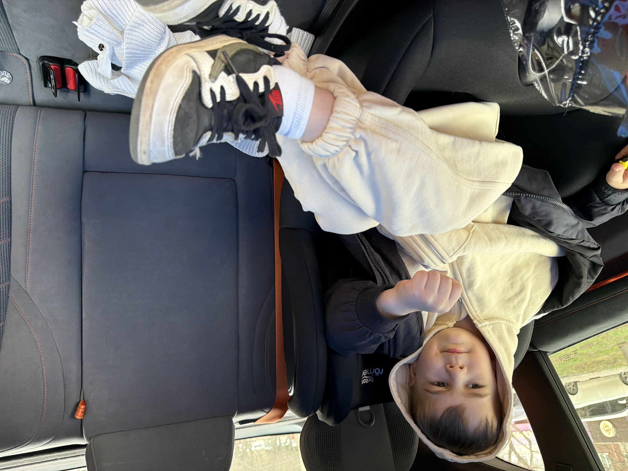 A child is in a car seat in the back of a vehicle. The child is wearing a hooded sweatshirt, a jacket, and white socks with sneakers on one foot resting on a white sweater. The car has gray upholstered seats. The location cannot be determined from the image.