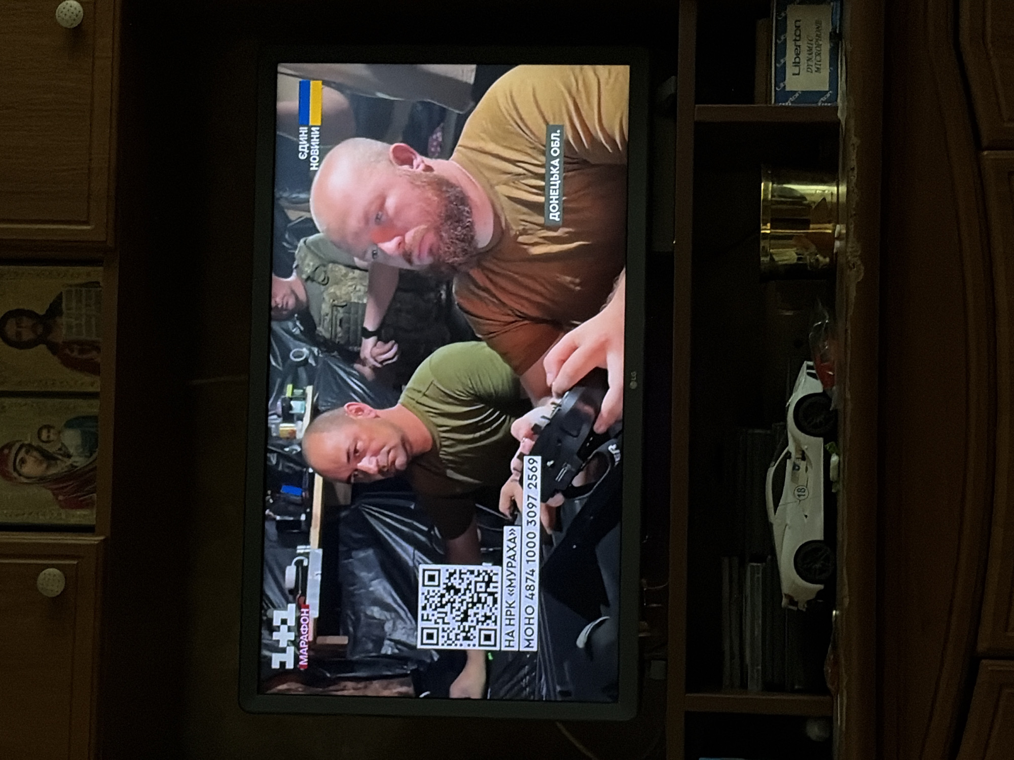 The image captures a television screen displaying a news broadcast. On the screen, two men are visible. The man on the left is wearing a green t-shirt, and the man on the right has a beard and is also wearing a brown t-shirt. The screen shows a QR code and text in Ukrainian, including "НА НРК «МУРАХА»" and "МОНО 4874 1000 3097 2569". A Ukrainian flag is also displayed next to the text "ЄДИHІ НОВИНИ". The broadcast appears to be from the channel "1+1 МАРАФОН". The text "ДОНЕЦЬКА ОБЛ." is visible in the bottom right corner of the screen, indicating the broadcast's geographical focus. The television is placed on a wooden shelf unit. Below the television, a white toy sports car with the number "18" on its side is visible, along with a gold-colored cylinder and a box for a "Liberton DYNAMIC MICROPHONE". The background of the television screen shows a dark, possibly makeshift, environment with black sheeting. The overall setting is indoors, with religious icons visible on the wall above the television.