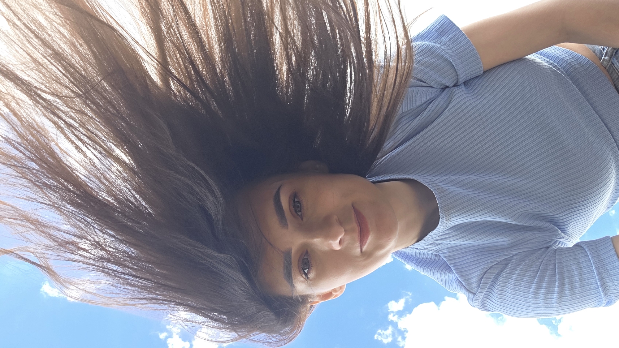 A young woman with long, dark brown hair is captured from a low angle against a bright blue sky with scattered white clouds. Her hair is being blown upwards and to the right by the wind, framing her face. She is wearing a light blue, ribbed short-sleeved t-shirt. Her gaze is directed upwards and slightly to the left, and she has a faint smile. The scene appears to be outdoors, possibly in a park or open area, given the sky and clouds. The location is identified as Дарьино, Russia.
