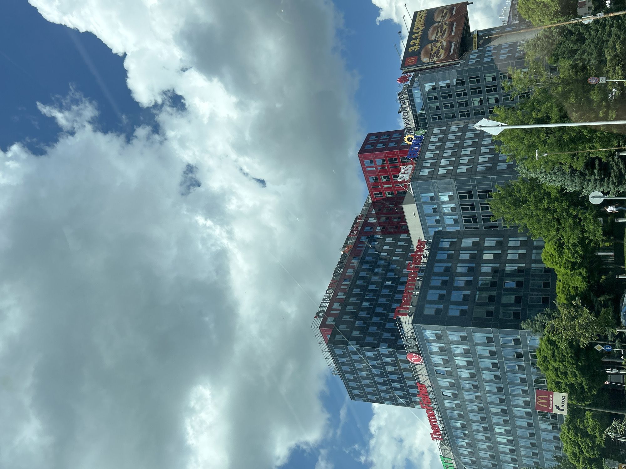 This image depicts a modern business park located in Sofia, Bulgaria, specifically identifiable as part of Business Park Sofia due to the visible corporate signs and Cyrillic text. The scene is dominated by multiple multi-story office buildings under a partly cloudy sky.
The upper portion of the image features a bright blue sky with extensive white and grey cumulus clouds. Faint contrails are visible crossing the sky.
In the mid-ground, a prominent dark-grey and blue-windowed office building stands on the left. Its facade displays large red illuminated signs for "ThermoFisher" and the "LG" logo. Below these, a smaller "ThermoFisher" sign is also visible. To its right and slightly behind, a taller building features red upper stories and displays "TINO" and "OZON" in red lettering on its roofline. Further to the right, another building, partially red, features "SGS" and "photomate" signs.
On the far right, a large billboard for McDonald's advertises "3.2.1.CHEESE" burgers. At a lower elevation on the left, a smaller McDonald's sign with "I'm lovin' it" includes a white arrow and Cyrillic text reading "ВХОД" (Entrance).
The lower section of the image is characterized by dense, vibrant green deciduous trees and some darker green coniferous trees, partially obscuring the base of the buildings and ground-level elements. Amidst the trees, a white street sign and a lamppost are visible. A single individual, dressed in a light-colored top and dark trousers, is visible walking on the far left at ground level. A dark vehicle is partially visible behind the trees on the bottom left. The overall lighting suggests a sunny daytime environment with diffuse light from the clouds.