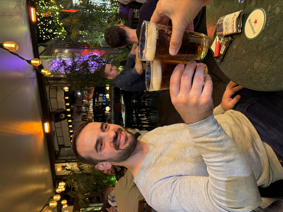 A man with a beard smiles while holding a beer in a busy outdoor bar. He is seated at a table with others, including one man holding a second beer in a toasting gesture. Overhead lights, including string lights and mounted lamps, illuminate the scene, while in the background, other patrons are seated. A Heineken coaster and cigarette paraphernalia sit on the table. The image is rotated ninety degrees to the left.
