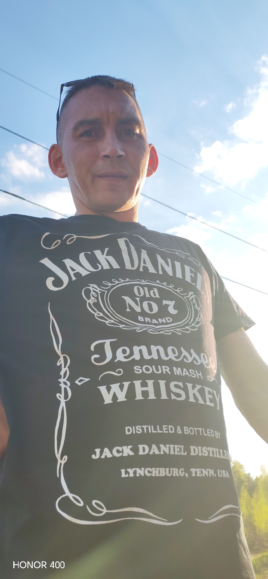 A man is posing outdoors, wearing a black t-shirt featuring the Jack Daniel's logo. The sky is visible in the background, partially obscured by bright sunlight and clouds. There are powerlines visible in the background. "HONOR 400" is written in the lower left corner. The location is Noviy, Russia.