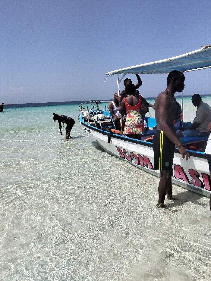 The image depicts a coastal scene in Wasini, Kenya, featuring a white motorboat with a blue interior and a light blue canopy, anchored in clear, shallow turquoise water under a clear blue sky. The boat, labeled "BOOM WASINI" in pink and blue lettering on its hull, contains at least five individuals of African descent.
In the immediate foreground, to the right of the boat, a shirtless male of African descent, wearing black shorts with yellow stripes, stands in the water, facing left in profile. Inside the boat, an adult female in an orange floral dress stands facing away from the viewer. Behind her, at least two males are visible; one wears a white tank top, and another holds onto the canopy support. Towards the stern, another male in a light-colored polo shirt is seated. A sixth male is partially visible in the boat, grasping the canopy frame.
To the left of the boat, a seventh individual, appearing as a dark silhouette, stands in the shallow water, bent over at the waist, reaching towards the surface of the water. The water is calm, revealing the sandy bottom beneath. A faint horizon line separates the ocean from the sky, and a small, dark vessel is visible in the far distance on the left.