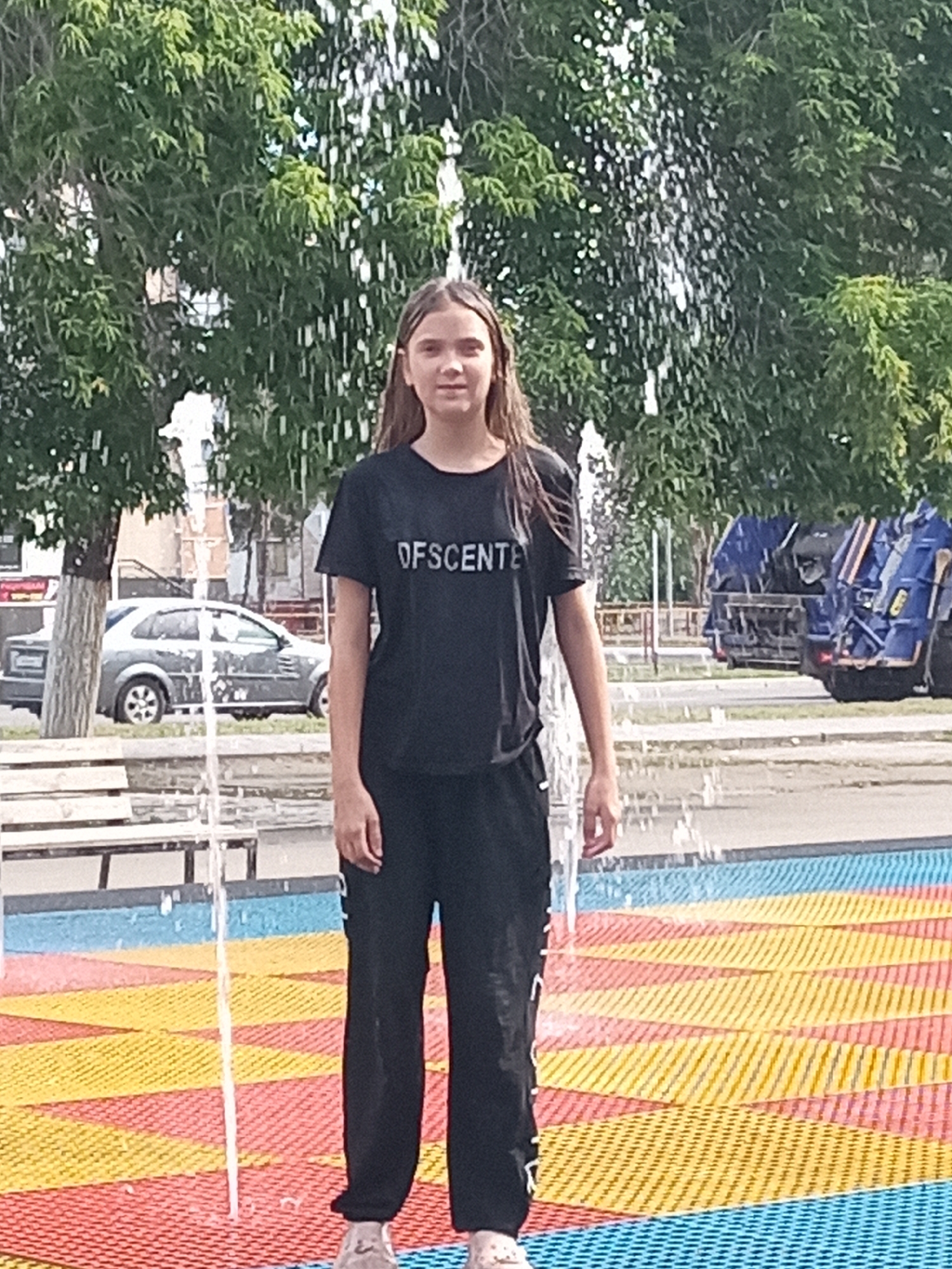 In Rudny, Kazakhstan, a young girl with long brown hair, wearing a black t-shirt with the text "OFSCENTE" and black pants, stands in a splash pad. Water sprays from a fountain above her, creating a curtain of water. She is positioned on a colorful, textured surface of red, yellow, and blue geometric patterns. In the background, trees line a street, with a silver car parked and two blue garbage trucks visible further down the road. A wooden bench can be seen to the left of the splash pad.