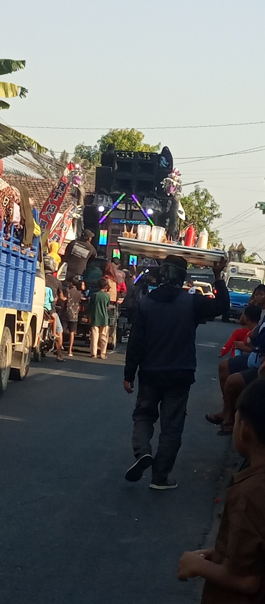 In Jekulo, Indonesia, a procession moves down a paved road under a clear sky. A man walks away from the camera, carrying a large, flat tray of food items, including bottles of condiments and what appear to be fried snacks, on his head. He is dressed in a dark hooded jacket and dark pants. Behind him, a large sound system is mounted on a vehicle, adorned with colorful LED lights and decorative elements, including what looks like a traditional puppet. Several people are visible around the sound system and in the background, including individuals standing in a blue truck on the left, and a crowd of onlookers gathered along the right side of the road. The environment suggests a festive or celebratory event, possibly a parade or street party, taking place during daylight hours.