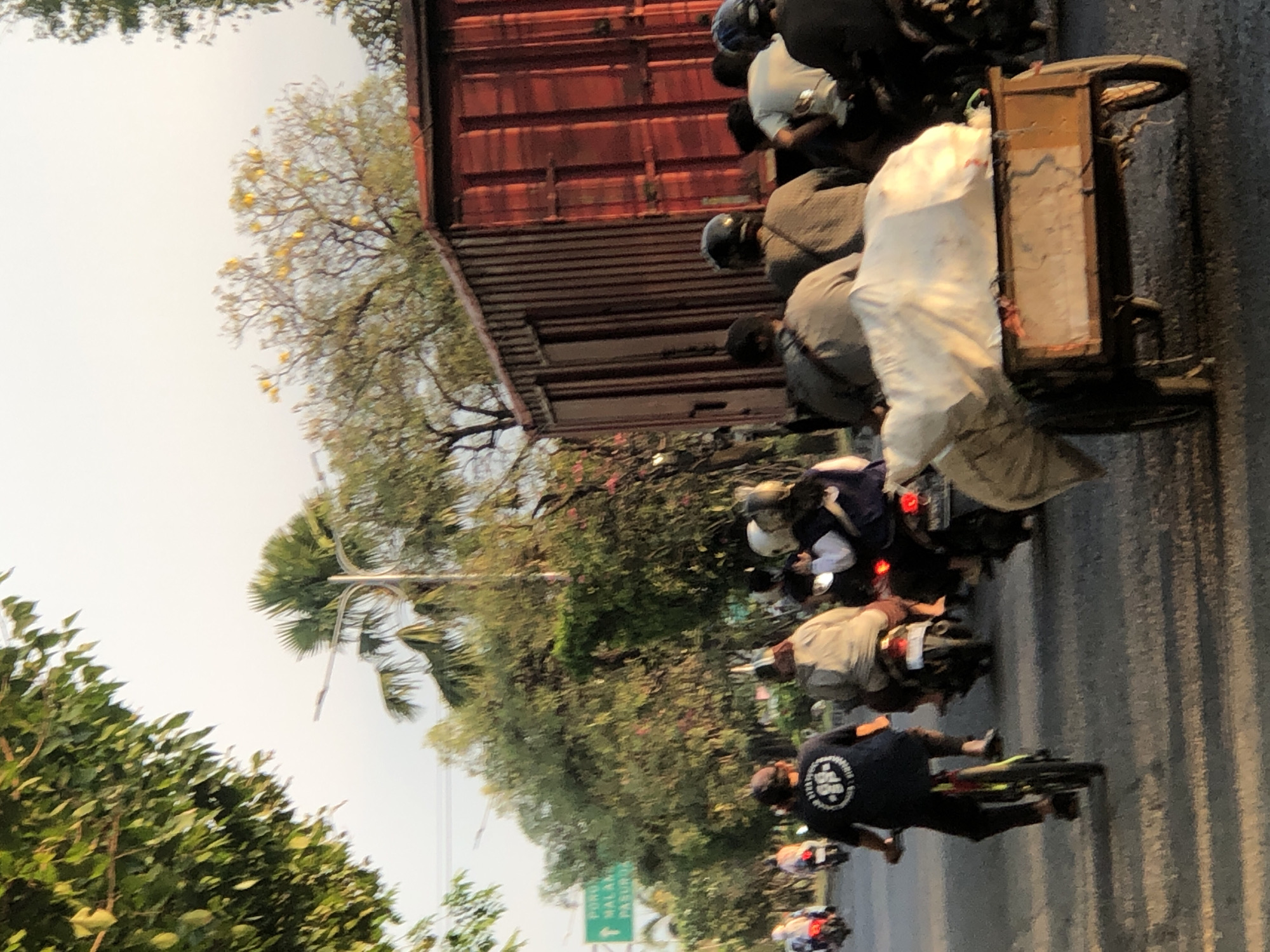 A group of people on motorcycles and a bicycle are traveling on a road in Gelam, Indonesia. A large, red shipping container is parked on the side of the road. In the foreground, a cart filled with a white tarp is being towed by a motorcycle. The scene appears to be a busy street with trees and a sign in the background.