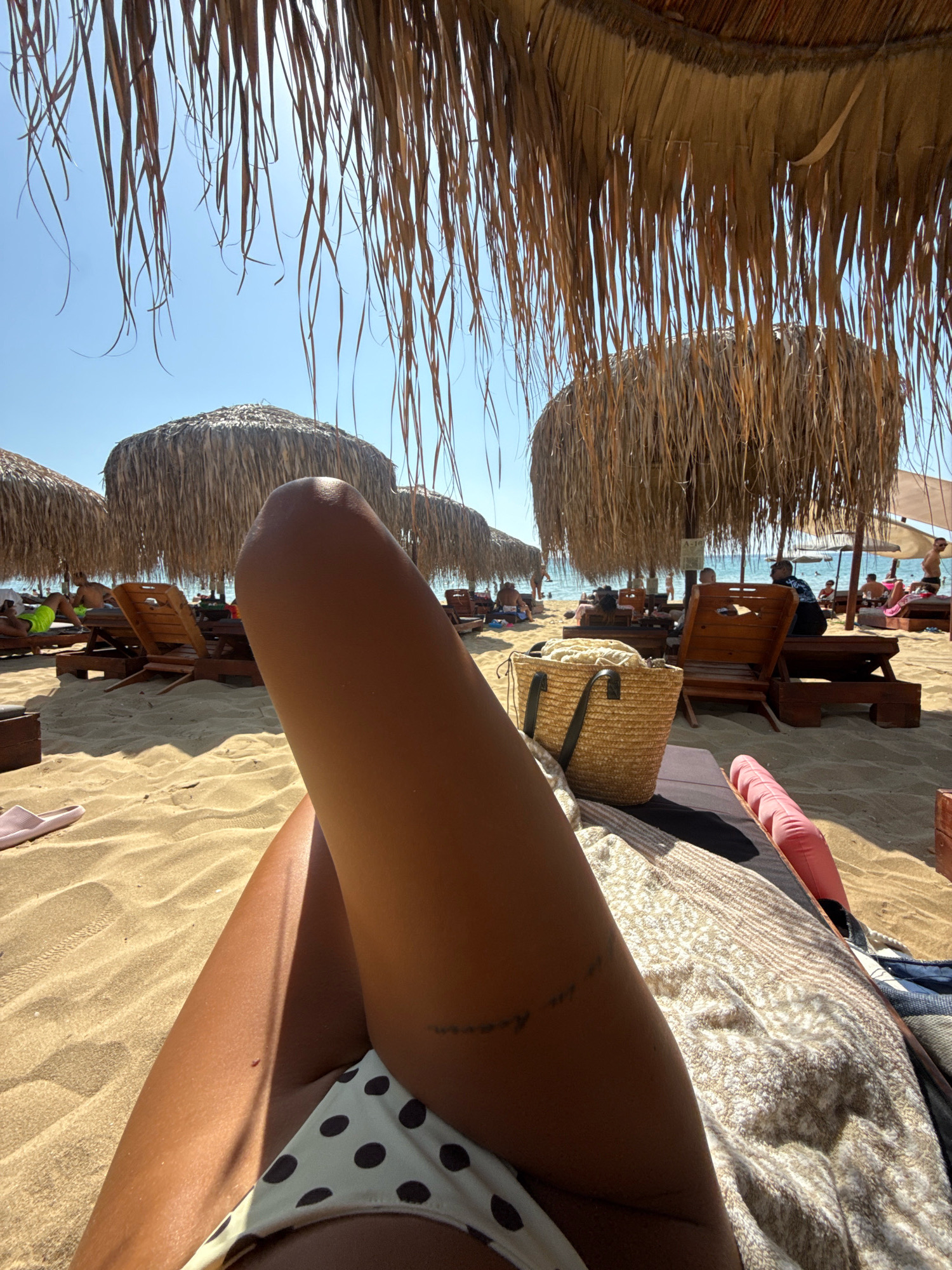 In Nea Peramos, Greece, this image captures a sun-drenched beach scene from a first-person perspective. The foreground is dominated by the legs and torso of an individual lying on a lounge chair. Their legs are tanned, and one leg has a faint tattoo. A white bikini bottom with dark polka dots is visible. A patterned blanket or towel covers part of the lounge chair. To the left of the foreground, a pink sandal rests on the sand. Behind the individual, the sandy beach stretches out, lined with rows of wooden lounge chairs and thatched umbrellas. Several other beachgoers are visible in the mid-ground and background, some sitting on chairs and others walking. The sea is visible in the distance under a clear blue sky. The overall impression is one of a relaxed day at the beach.