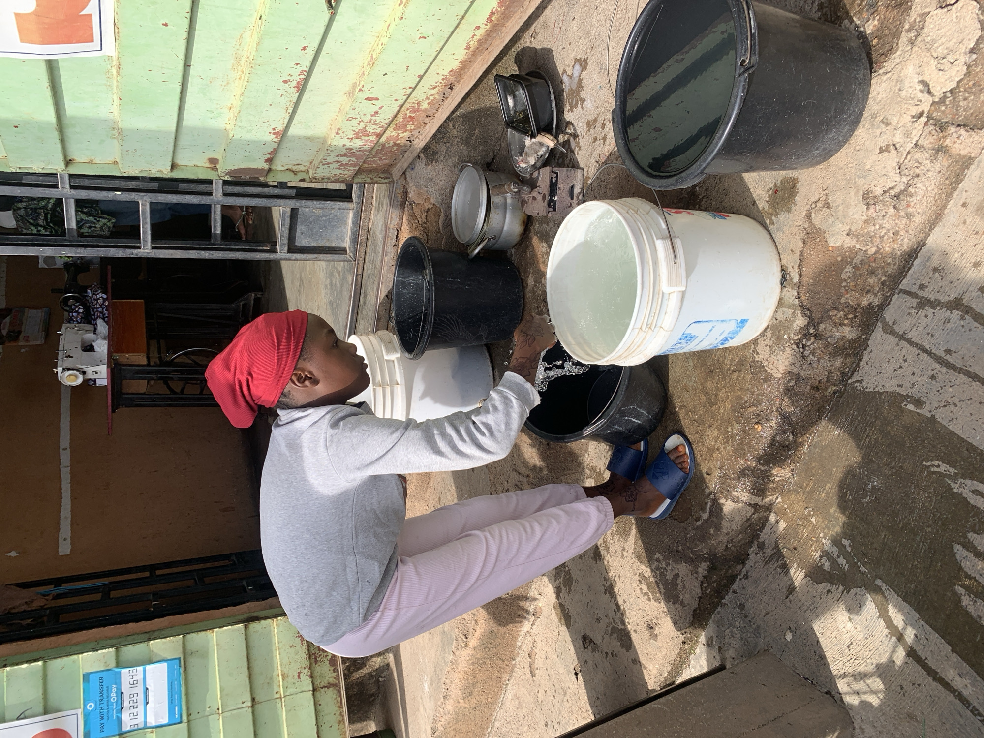 This image depicts a person engaged in the task of transferring water between buckets, likely outside a small business or residence in Jos, Nigeria, given the visible signage and context. The scene is brightly lit by strong sunlight, casting distinct shadows on the ground.
The primary subject is a person, appearing to be young, who is kneeling or squatting on a rough concrete surface. They are wearing a red head covering, a light grey long-sleeved top, and light pink pants. Intricate dark patterns, possibly henna or tattoos, are visible on their hands and feet, which are clad in blue and white slide sandals. The person is actively pouring clear water from a large white plastic bucket into a black plastic bucket placed on the ground directly in front of them.
Several other items are scattered around the work area: another large black bucket, partially filled with water, sits further back to the right. A smaller white bucket is near the person's left. On the ground behind the buckets, various metal household items are visible, including a lidded pot and a small, possibly antique, iron. The concrete ground is visibly wet from spilled water.
The background features a weathered building with a facade made of light green corrugated metal sheets, heavily marked by rust. An open doorway, partially covered by horizontal metal bars, provides a glimpse into the interior, where a sewing machine is discernible, suggesting the space might be a tailoring shop or similar establishment. Two signs are affixed to the green wall: one with obscured red text, and another blue sign prominently displaying "PAY WITH TRANSFER" for "OPay," along with the phone number "0812 229 1643." This transfer of water appears to be a routine daily chore.