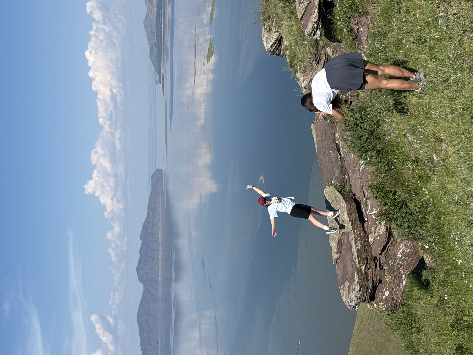 In the foreground, a person in a red cap, white t-shirt, and black shorts stands on a rocky outcrop overlooking a large body of water. They are posing with arms outstretched, as if in mid-leap or dance. In the mid-ground, another person, seen from behind, is kneeling and appears to be taking a photo with a smartphone. They are wearing a white t-shirt and a dark skirt. The environment consists of a grassy hillside dotted with wildflowers leading to the rocky ledge. Beyond the ledge, a vast, calm body of water stretches towards distant, hazy mountains under a bright blue sky with scattered white clouds. The water reflects the sky and the mountains, creating a serene and expansive landscape. This scene appears to be located in the vicinity of Ust'-Abakan, Russia, a region known for its natural beauty and large water bodies.