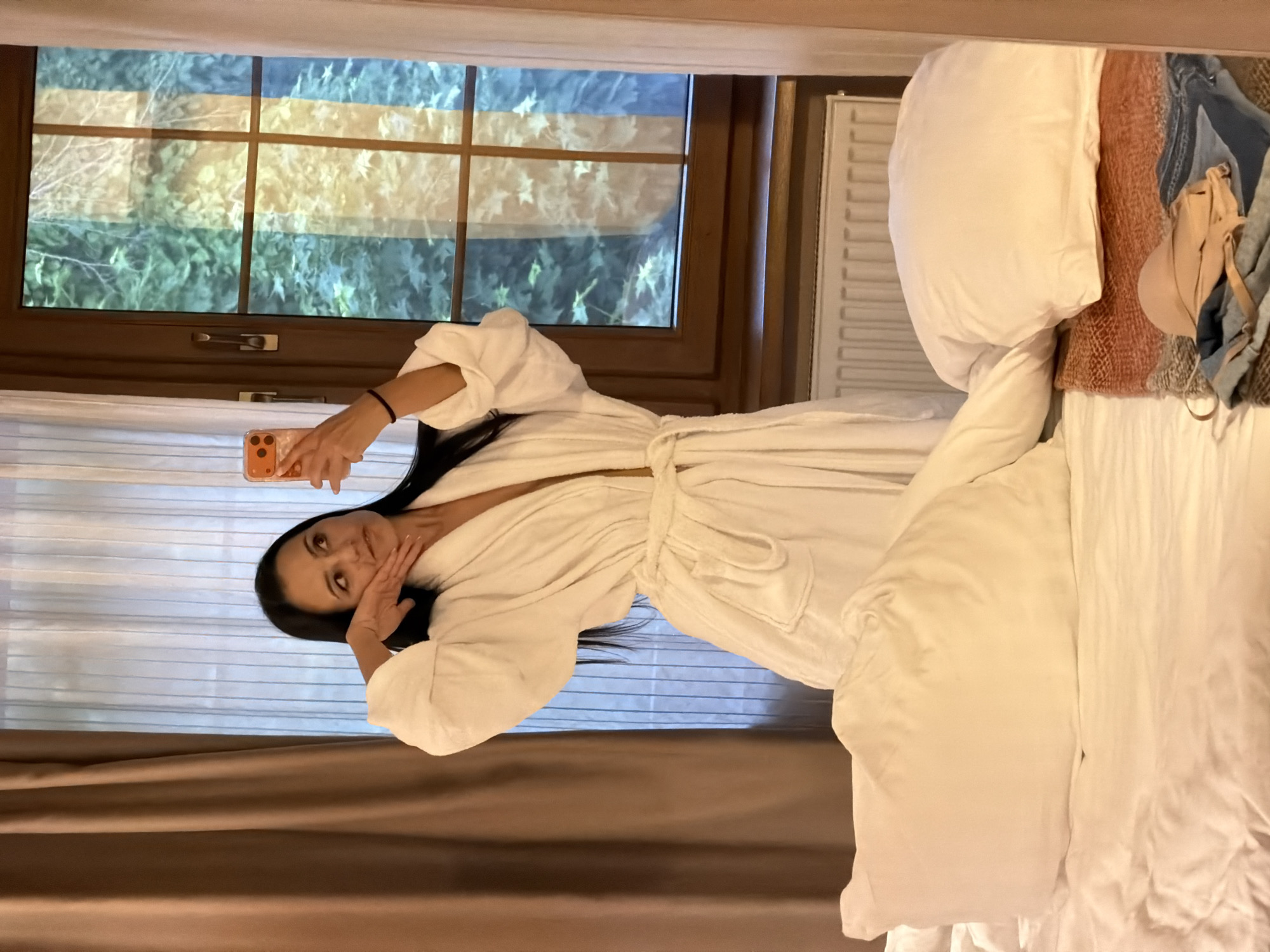 An adult female is captured in a mirror selfie inside a room, presumably in Svetlogorsk, Russia. She stands center-frame, dressed in a white terrycloth bathrobe tied at the waist. Her long, dark hair is down. She holds an orange smartphone with a triple-lens camera in her right hand, positioned to take the photograph. Her left hand is raised to her jawline, fingers splayed, and her gaze is directed towards the upper right, away from the camera lens. A black hair band is visible on her right wrist.
The background features a window with a dark brown wooden frame, divided into multiple panes, revealing lush green foliage outside. Vertical reflections or distortions, including bands of yellow and blue, are present on the window glass. To the left of the window, a heavy brown curtain is partially drawn, while a lighter, possibly sheer, curtain is visible on the far right. A white horizontal radiator is located directly beneath the window.
In the foreground, the lower section of a bed is visible, covered with white sheets and multiple white pillows. On the right side of the bed, a reddish-orange textured blanket or throw is laid out, with blue denim fabric (possibly jeans) underneath and a light beige bra resting on top.