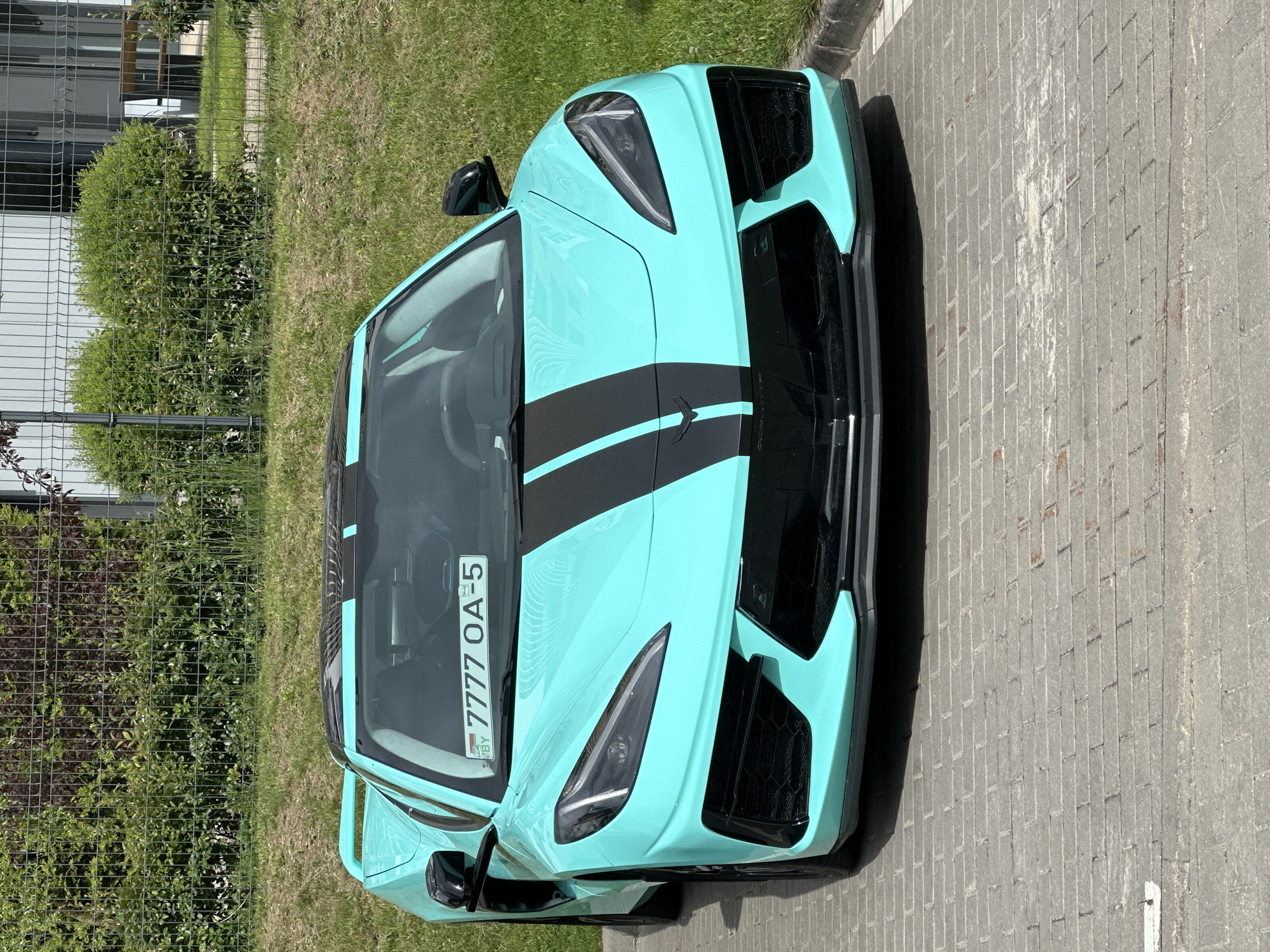 Observed outdoors in Бараўлянскі сельскі Савет, Belarus, is a light blue sports car with black racing stripes. The vehicle is parked on a brick surface adjacent to a grassy area bordered by a wire fence. The license plate reads "7777 OA-5" and includes "BY," the country code for Belarus. No people are visible.