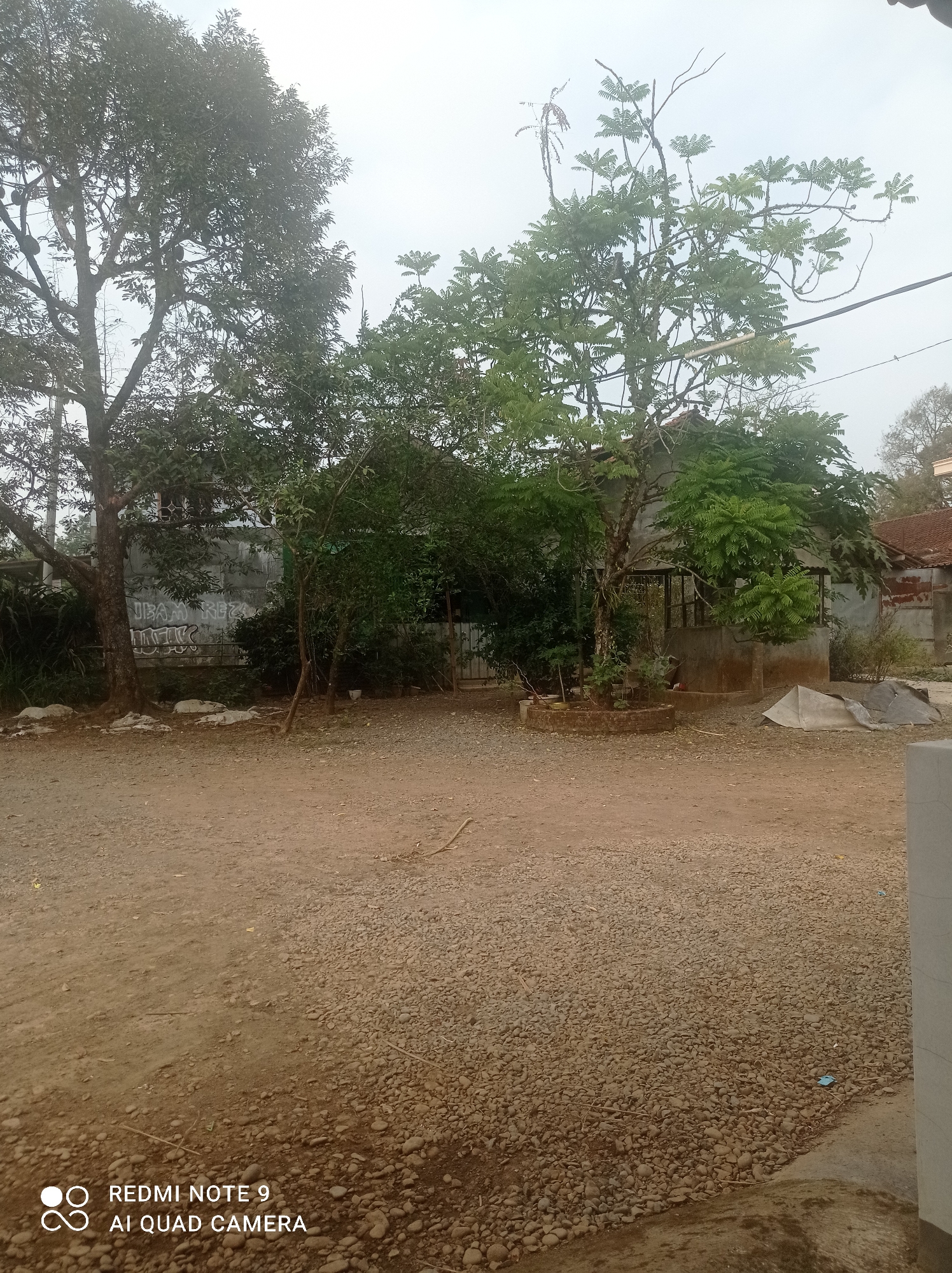 The image shows an outdoor scene with a dirt and gravel ground, several trees, and a simple building in the background. The trees are lush with green leaves, and some appear to be quite old and large. The building in the background is made of concrete and has a tiled roof. There is some graffiti on the wall of the building. The sky is overcast, suggesting it might be a cloudy or rainy day. The overall impression is one of a rural or semi-rural area in a tropical climate.
The image is from Cipawon, Indonesia.