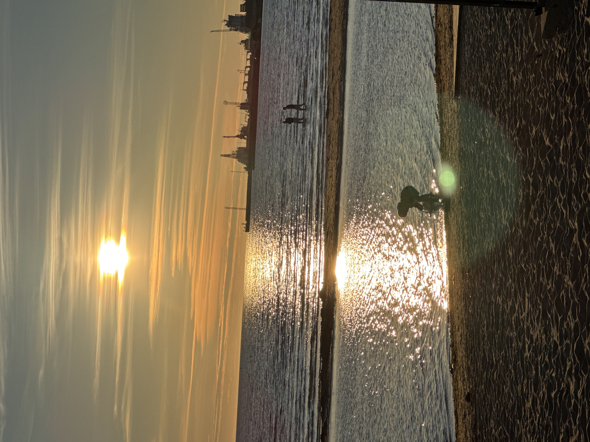 The image depicts a coastal scene at sunset, likely in Tallinn, Estonia, given the presence of industrial port structures on the horizon. The sun, a bright orb reflecting on the water, casts a warm, golden glow across the sky and sea. In the foreground, a sandy beach transitions into shallow water where a solitary figure, silhouetted against the light, appears to be playing or collecting something. Further out in the sea, two more figures are wading, their forms also indistinct against the glare. The middle ground is dominated by the vast expanse of the Baltic Sea, its surface rippled by gentle waves and shimmering with the sun's reflection. In the distance, a line of cranes and ship hulls of an industrial port breaks the horizon, suggesting a working harbor. The sky is a gradient of warm oranges and yellows near the horizon, fading into cooler blues and grays higher up, with wispy clouds streaking across. The overall atmosphere is tranquil and serene, capturing the end of a day by the sea.