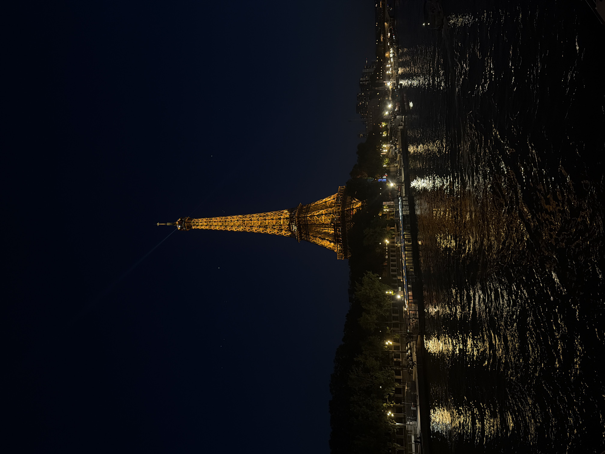 The image depicts the Eiffel Tower in Paris, France, illuminated at night. The tower stands prominently on the left, with its golden glow contrasting against the dark blue sky. The Seine River occupies the right side of the image, reflecting the lights from the city and the tower, creating shimmering patterns on the water's surface. Along the riverbank, streetlights and building lights cast a soft, warm illumination on the trees and structures, adding depth to the scene. There are no people visible in the image. The environment appears to be calm and serene, with the lights creating a tranquil ambiance.